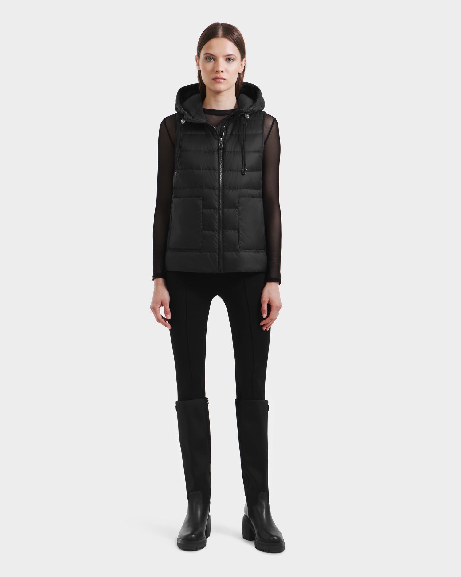 A woman faces forward wearing the Rudsak BETHANY - 8125625 Black modular hooded puffer vest over a sheer black long-sleeve top, fitted pants, and knee-high boots against a plain white background.
