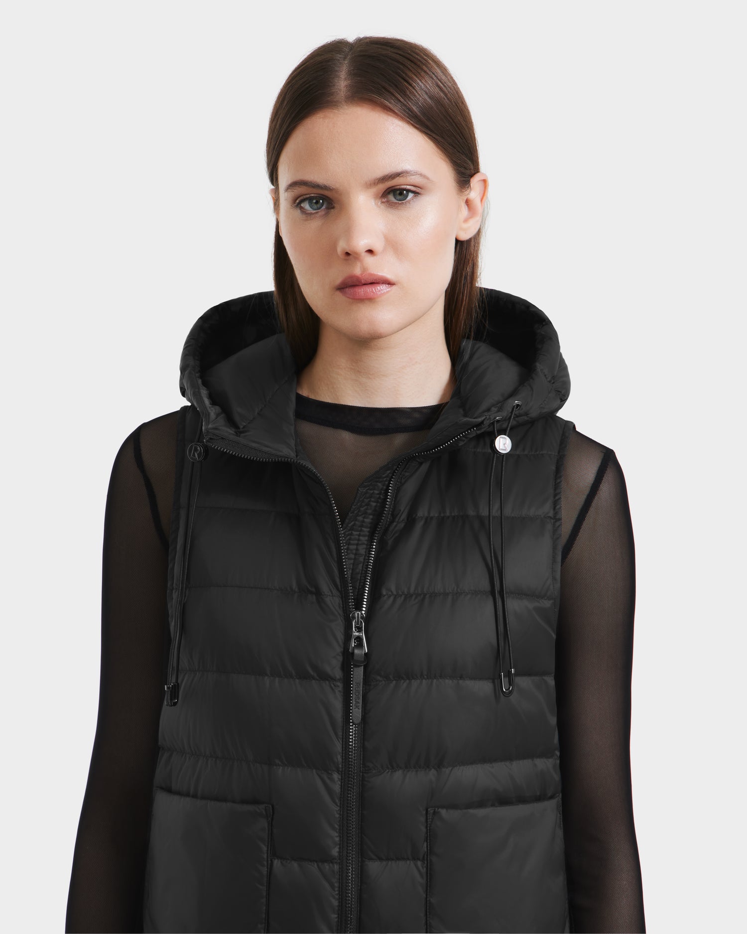 A woman with straight brown hair wears the Rudsak BETHANY - 8125625 Black modular puffer vest over a sheer black long-sleeve top, standing against a light gray background.