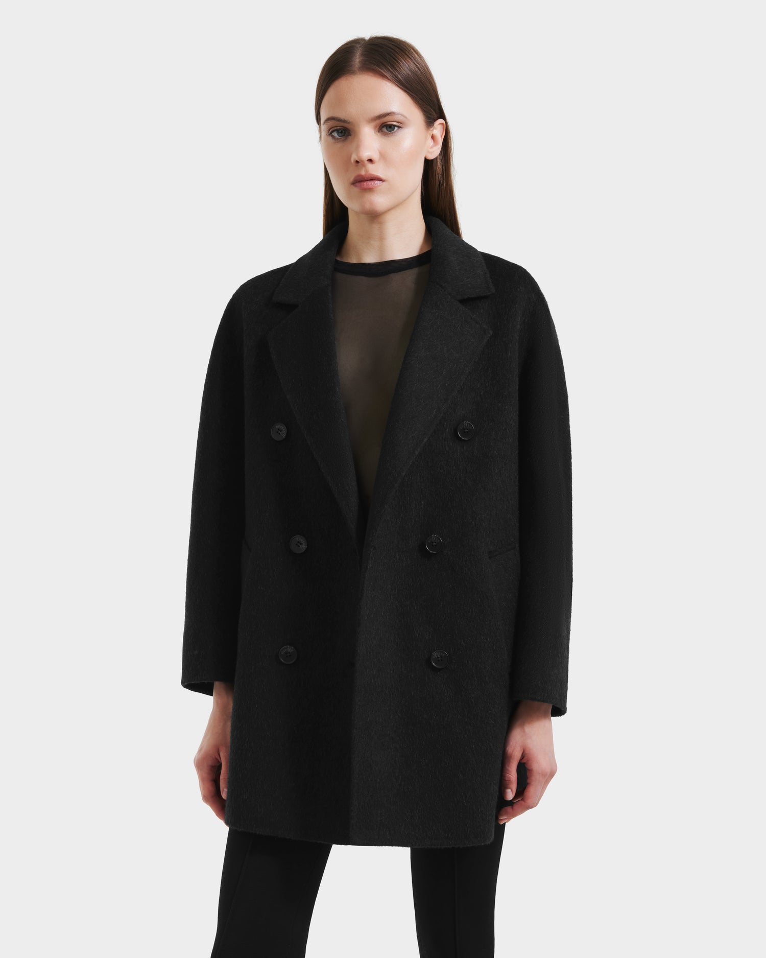 A woman with straight brown hair models the Rudsak BETHANY - 8125625 Black coat in a 3-in-1 silhouette over a sheer black top and black pants, standing against a plain white background.