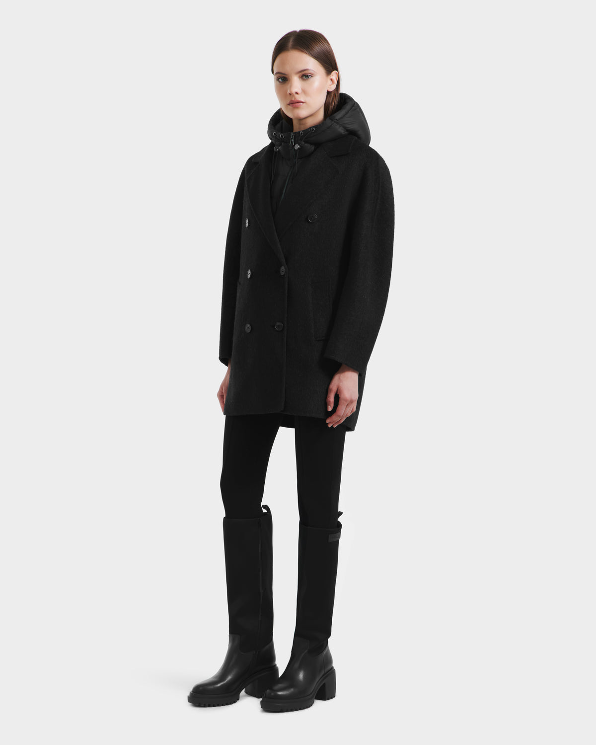 A woman faces forward wearing the Rudsak BETHANY - 8125625 Black coat over a hooded top, black pants, and knee-high black boots against a plain white background.