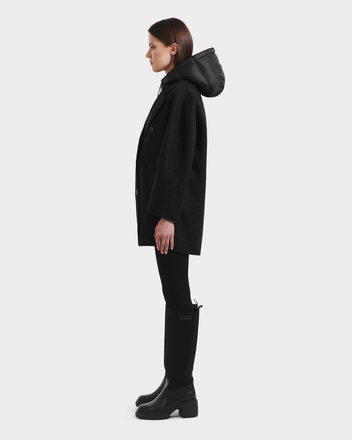A person stands in profile against a light background, wearing the Rudsak BETHANY - 8125625 Black 3-in-1 hooded coat, styled with black pants and knee-high boots.