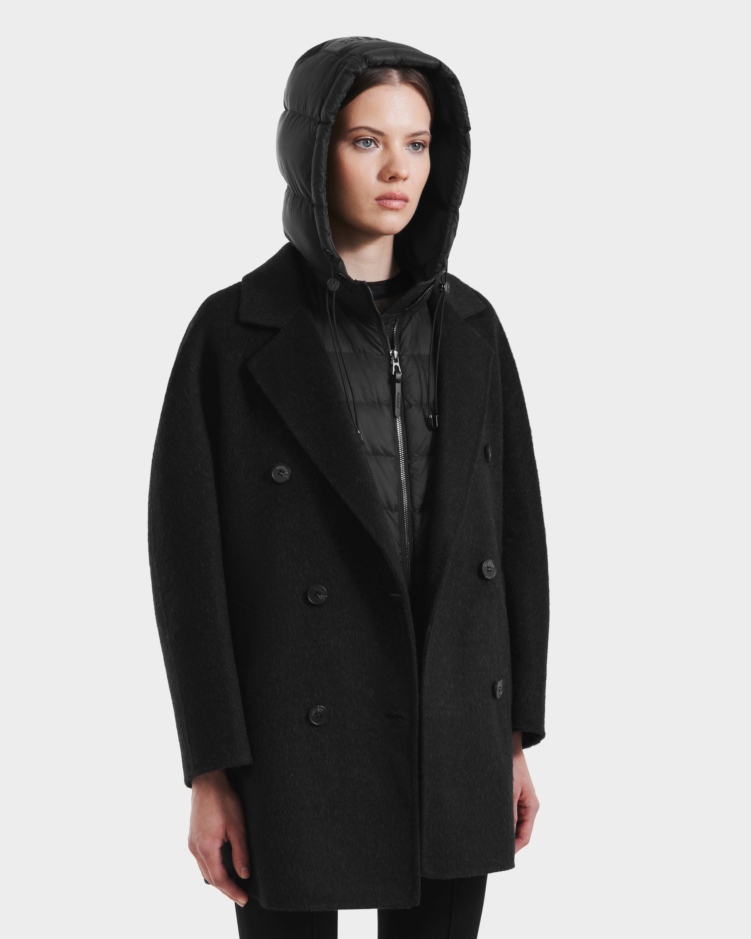 Someone wearing the Rudsak BETHANY - 8125625 Black modular outerwear—a black double-breasted wool-silk-yak coat layered over a black hooded puffer—stands against a plain white background.