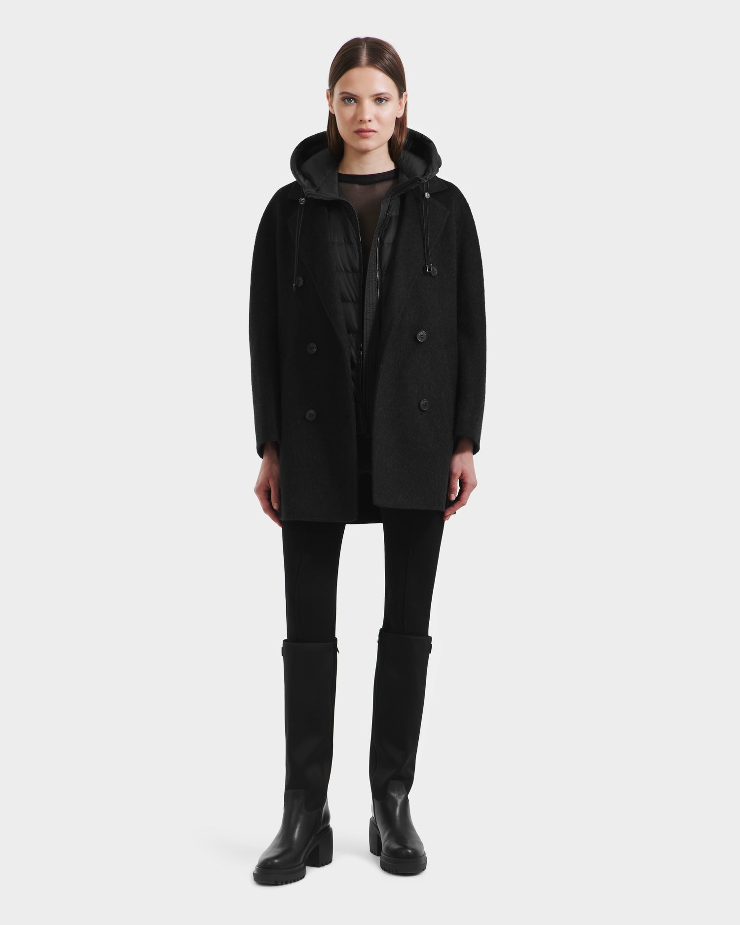 A woman faces forward, wearing the Rudsak BETHANY - 8125625 Black 3-in-1 coat with black leggings and knee-high boots, set against a plain light gray background.