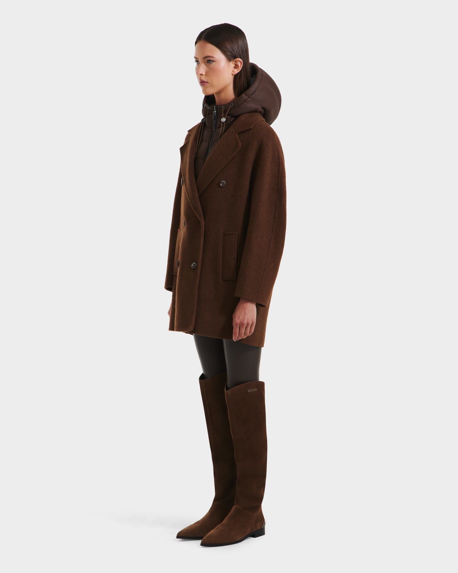 A woman wears the Rudsak BETHANY - 8125625 Mocha double-breasted coat with a hood, paired with black leggings and knee-high brown suede boots, standing against a plain white background.