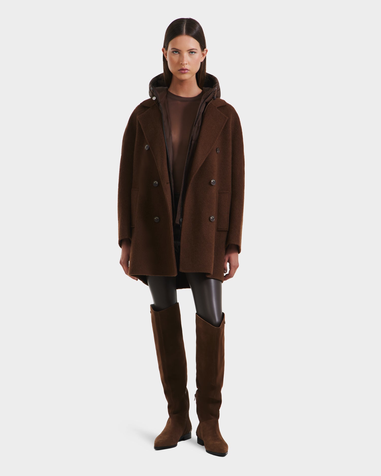 A woman faces forward in a Rudsak BETHANY - 8125625 Mocha double-breasted coat, paired with brown knee-high boots, black leggings, and a sheer brown top, set against a plain white background.
