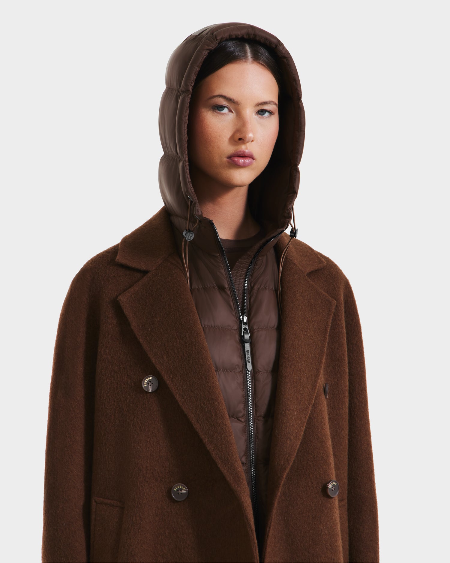 A woman wears the Rudsak BETHANY - 8125625 Mocha coat, layered over a glossy brown puffer with the hood up, presenting a sophisticated 3-in-1 look against a plain white background.