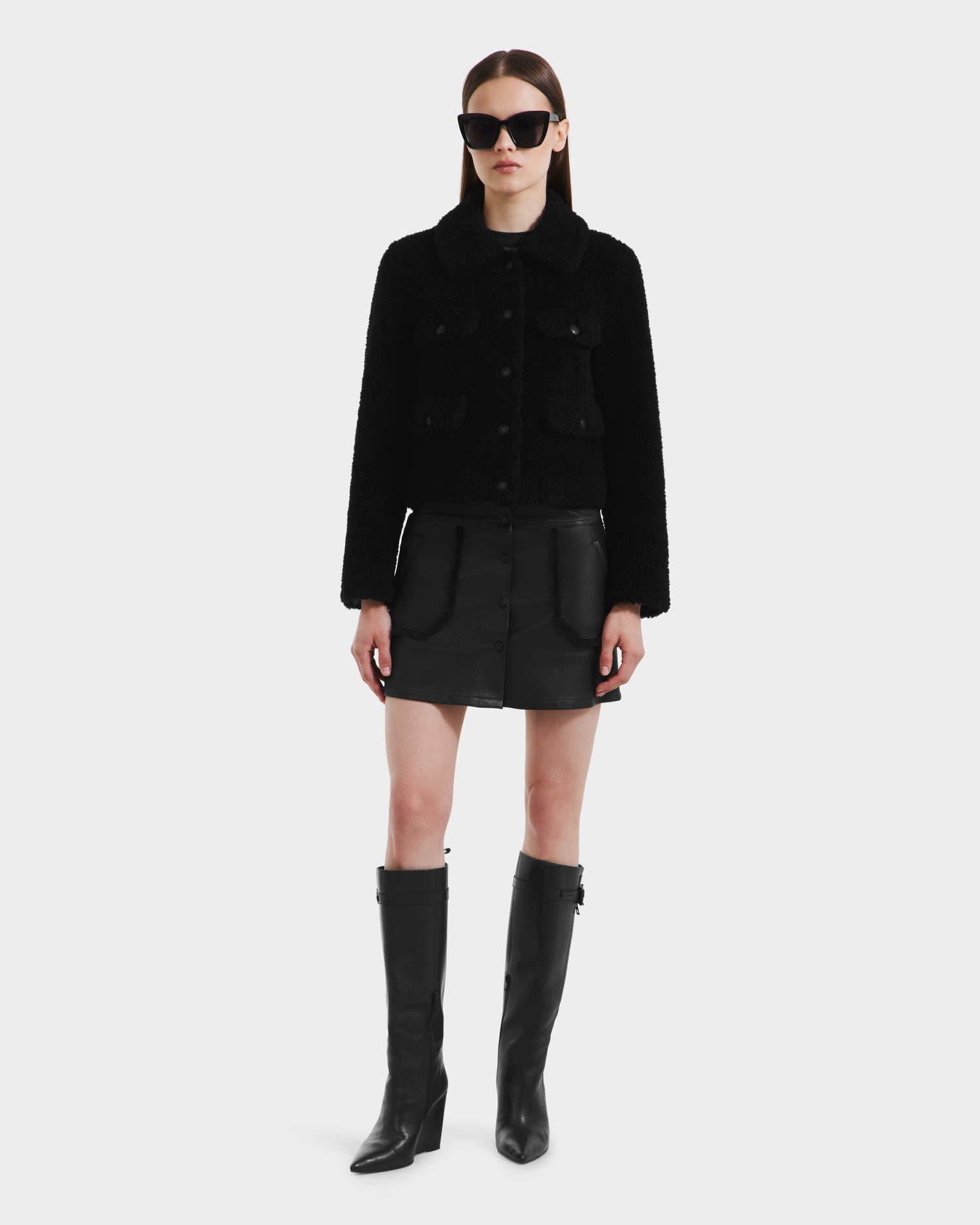 A woman faces forward in a Rudsak BLAIR - 8125513 Black women’s cropped teddy jacket, paired with a black leather mini skirt, knee-high boots, and large sunglasses against a white background for a sleek modern look.