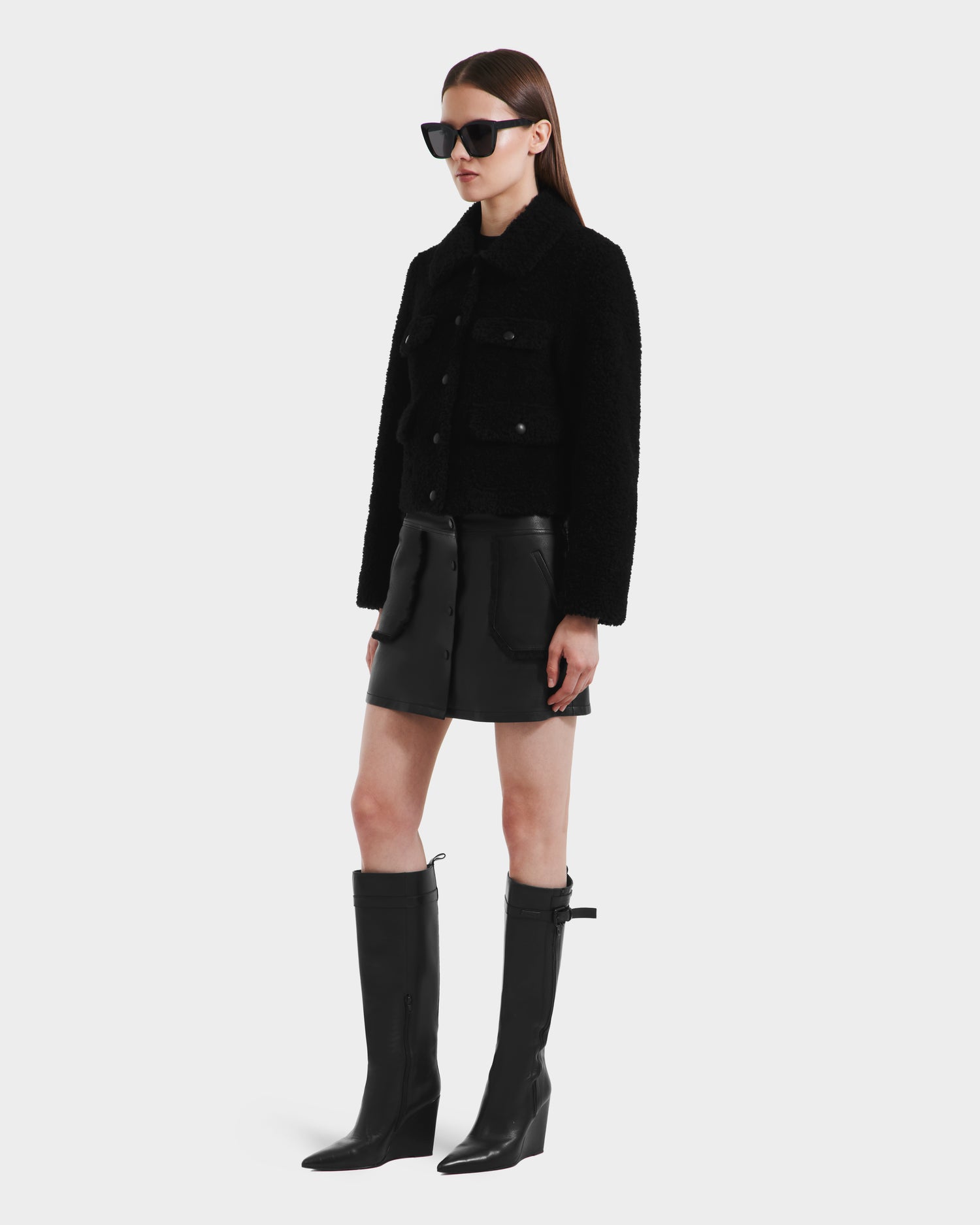 A woman wearing Rudsak’s BLAIR - 8125513 Black faux shearling jacket, paired with black sunglasses, a buttoned black leather skirt, and knee-high boots, stands against a plain white background.