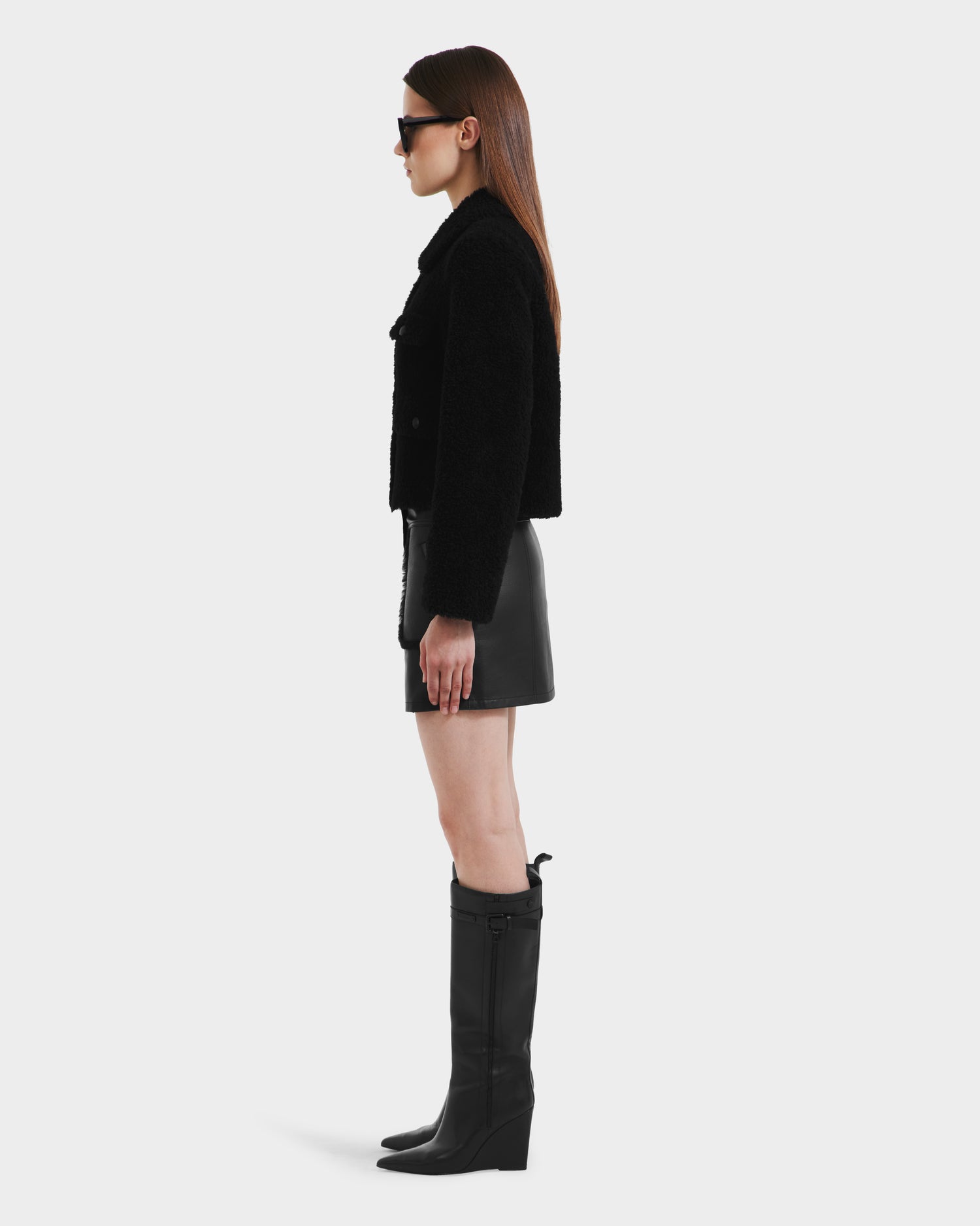 A woman stands in profile against a white background wearing the Rudsak BLAIR - 8125513 Black faux shearling jacket, a black leather mini skirt, tall black boots, and sunglasses.