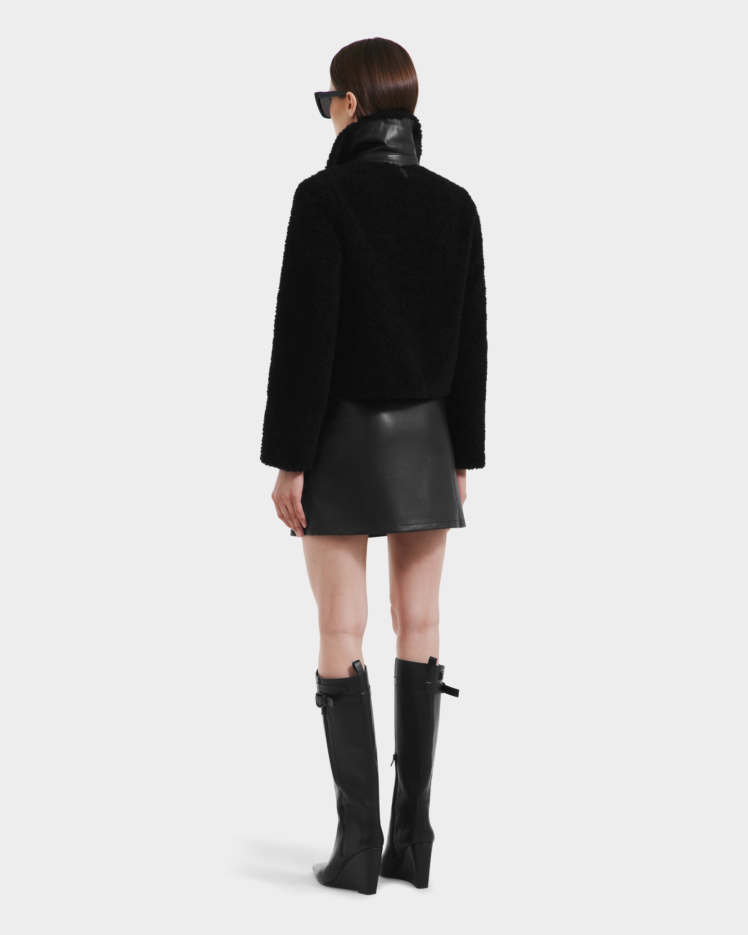 A woman with straight brown hair faces away from the camera in modern style, wearing the Rudsak BLAIR - 8125513 Black faux shearling jacket, paired with a black leather mini skirt, knee-high boots, and sunglasses against a white background.