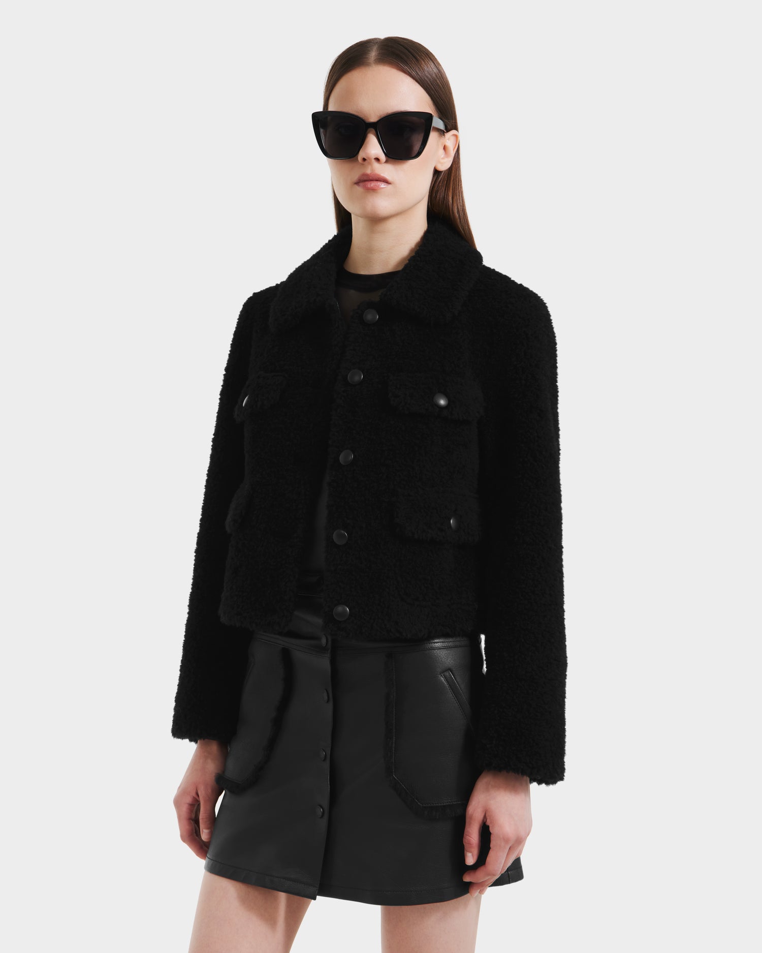 A woman wearing Rudsaks BLAIR - 8125513 Black womens cropped teddy jacket with textured front pockets, paired with black sunglasses and a black mini skirt, stands against a plain light background.