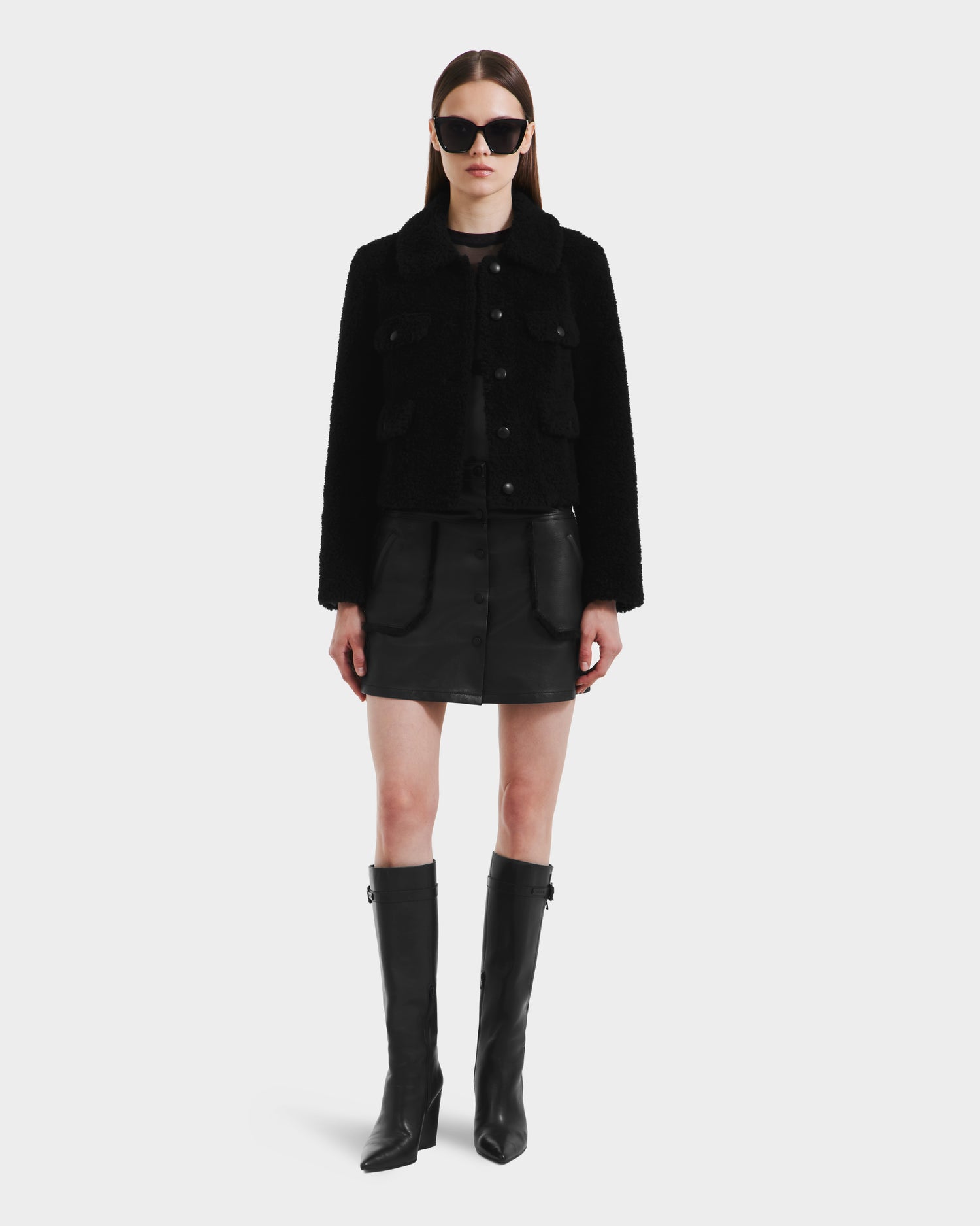 A woman stands facing forward in the Rudsak BLAIR - 8125513 Black cropped teddy jacket, black mini skirt, knee-high boots, and sunglasses, showcasing modern style against a plain light background.