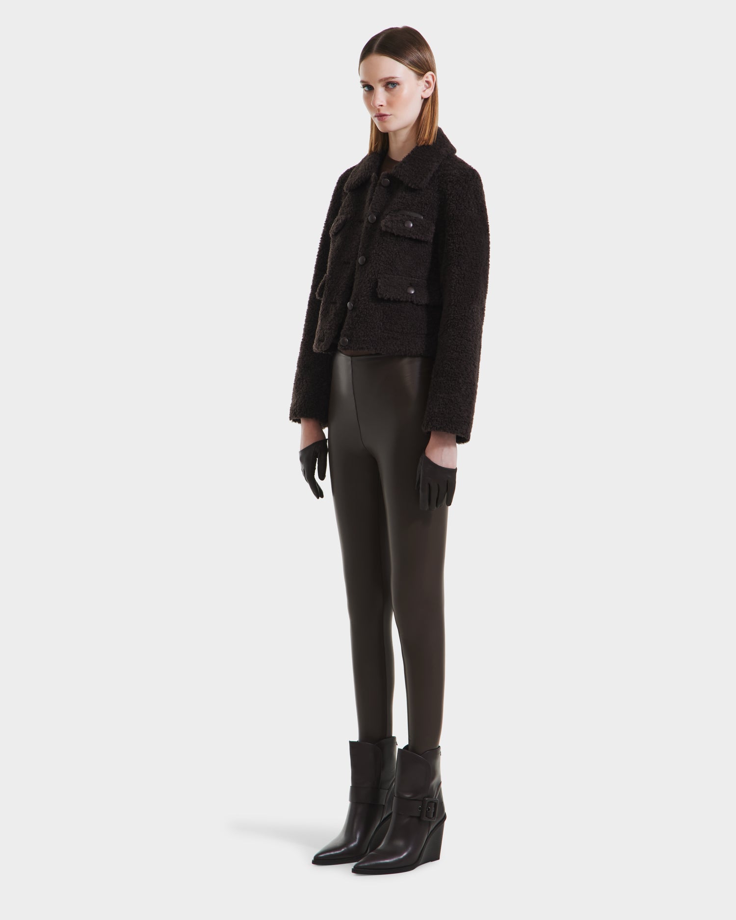 A woman with straight hair and a neutral expression stands against a plain background, modeling the Rudsak BLAIR - 8125513 Espresso womens cropped teddy jacket, styled with fitted dark pants, black ankle boots, and gloves.