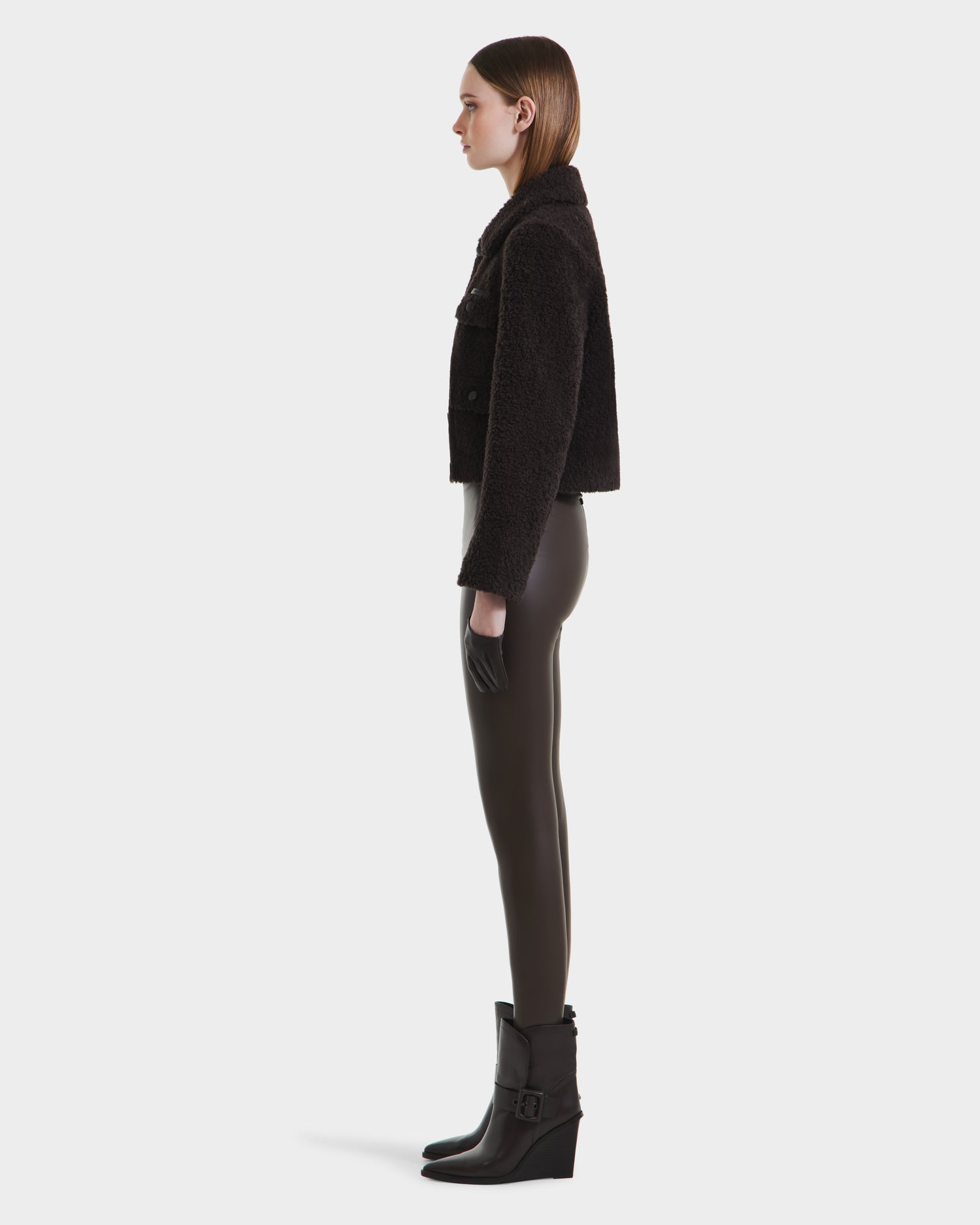 A woman in profile against a white background wears the Rudsak BLAIR - 8125513 Espresso womens cropped teddy jacket, black fitted pants, and modern ankle boots with buckles. Her straight brown hair is tucked behind her ears.