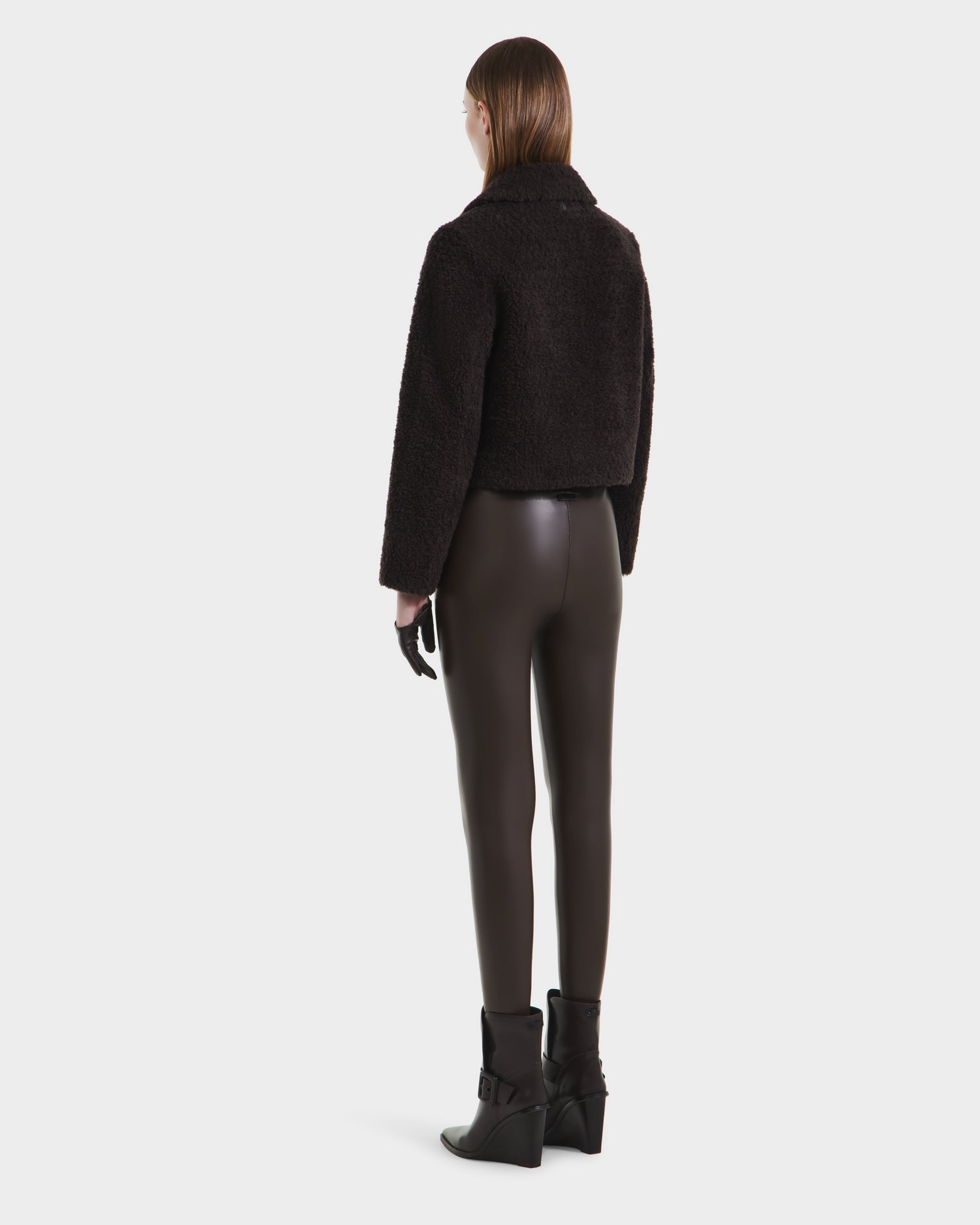 A woman with straight brown hair faces away, modeling the Rudsak BLAIR - 8125513 Espresso womens cropped teddy jacket with black leggings, gloves, and ankle boots, exuding modern style against a plain white background.