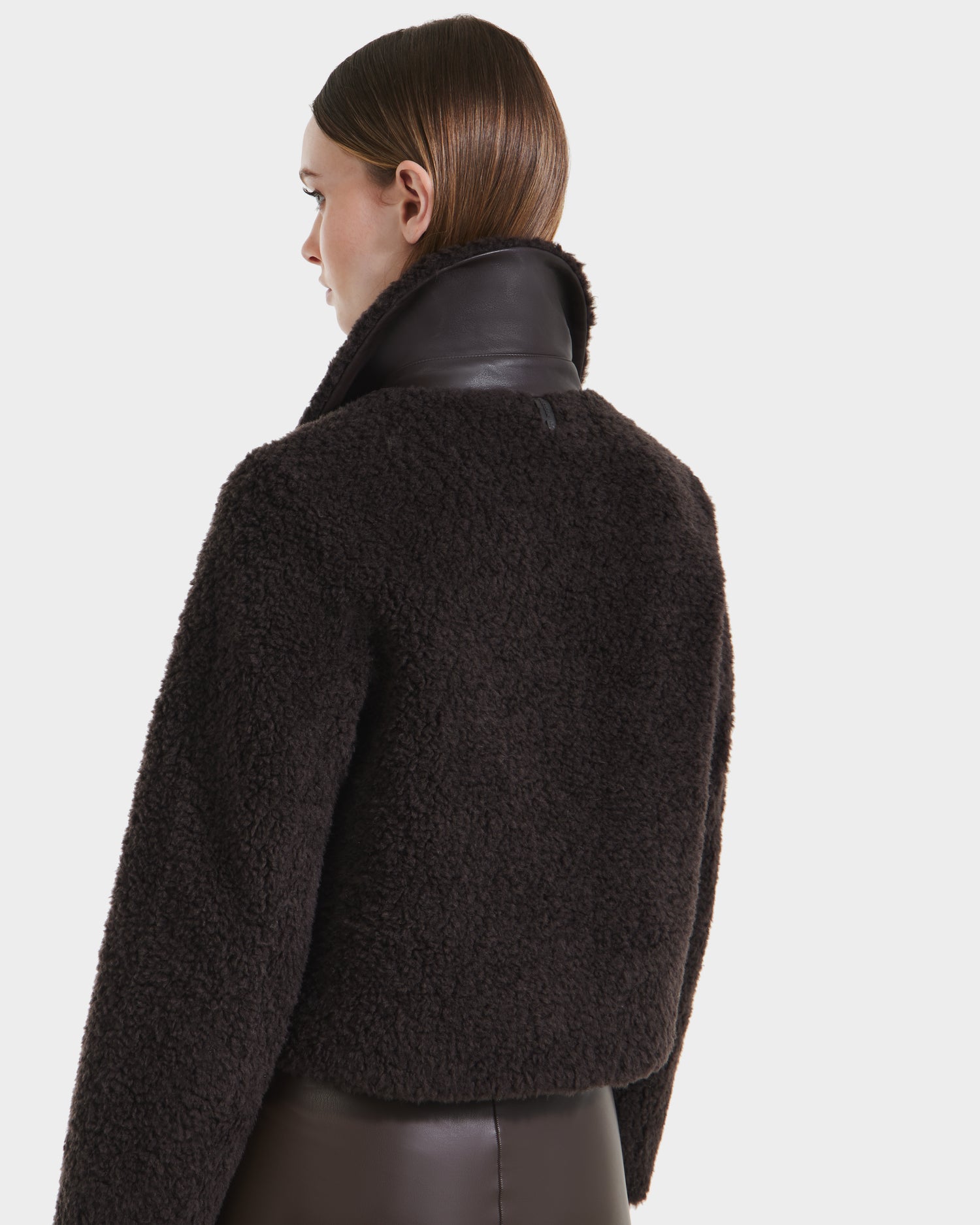 A woman with straight brown hair wears the Rudsak BLAIR - 8125513 Espresso cropped teddy jacket, featuring dark textured faux shearling and a high leather collar, shown from the back on a plain white background.