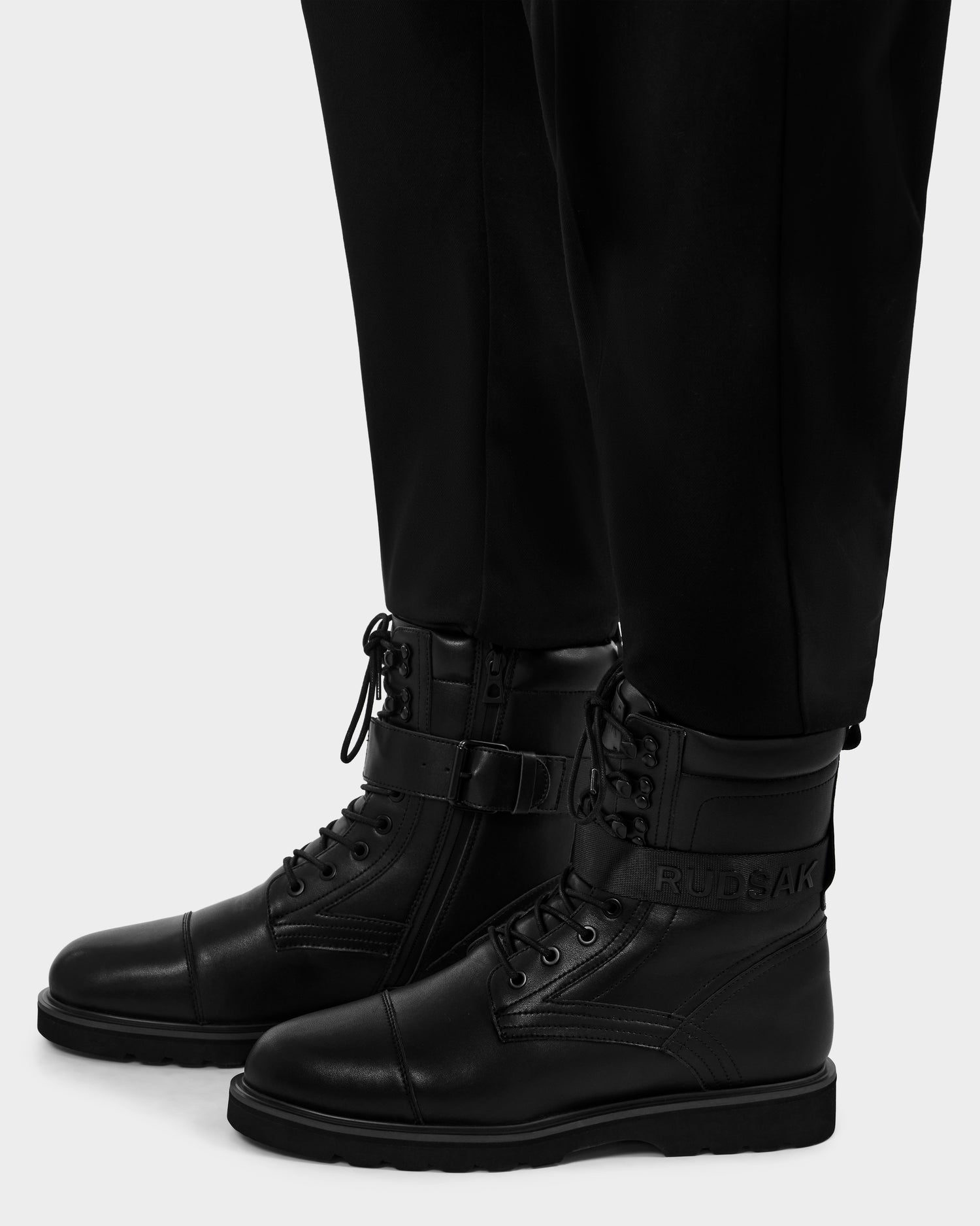 A person wears black pants and BLITZ - M223500 Black military-inspired leather lace-up boots with side zippers and a strap featuring the Rudsak brand name in raised letters.