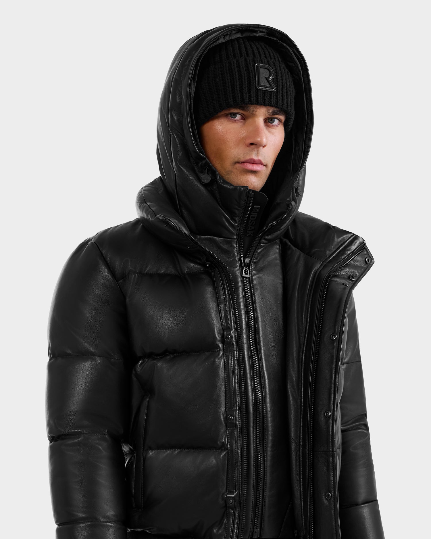 A person in the RUDSAK BOND X - 6122525 BLACK men’s winter jacket with RDS-certified down, hood, and a black knit beanie stands against a light background, looking to the side with a neutral expression.