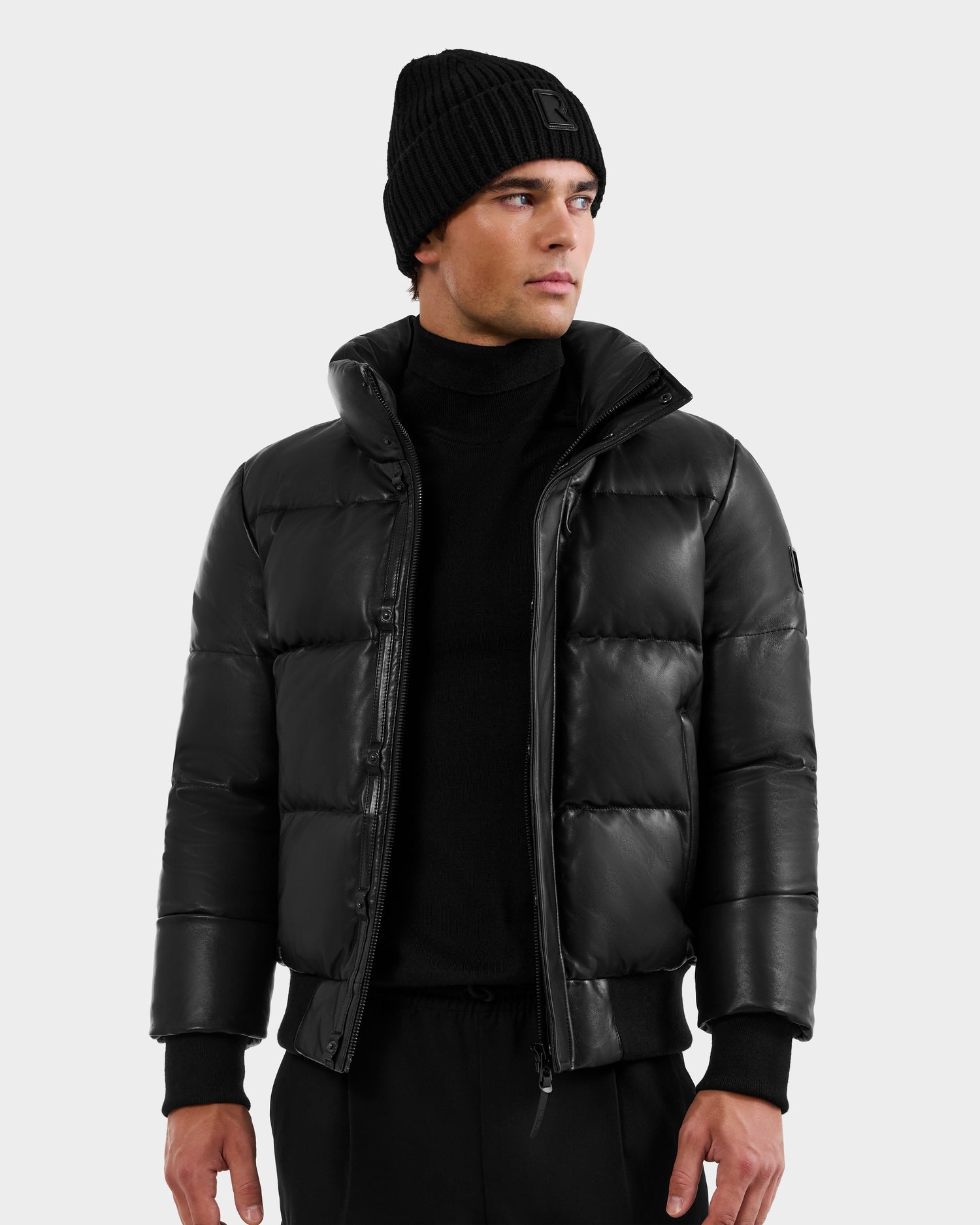 A man in the RUDSAK BOND X - 6122525 BLACK men's winter jacket—a sleek black puffer with RDS-certified down—stands against a plain light background, looking to his left with a neutral expression.