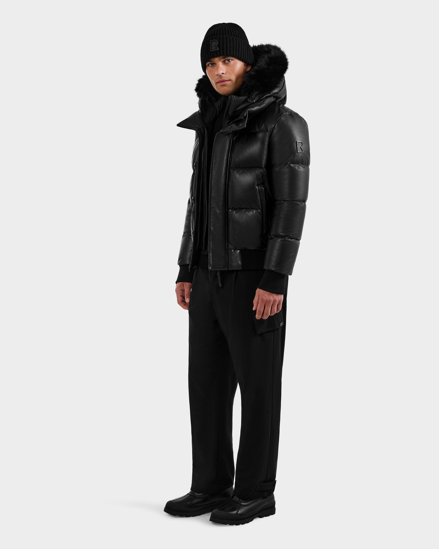 A person wearing the RUDSAK BOND X - 6122525 BLACK men’s winter jacket with RDS-certified down fill, black pants, and black shoes stands facing forward against a plain light gray background.