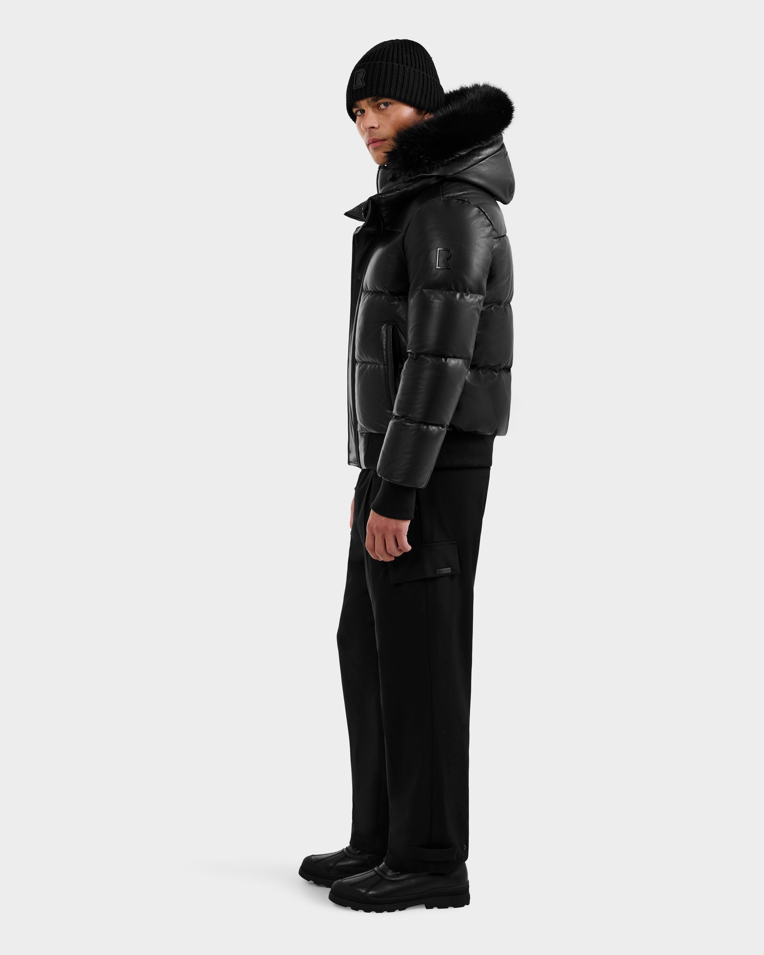 A person wearing the RUDSAK BOND X - 6122525 BLACK men's shiny black puffer jacket with RDS-certified down fill, a black knit beanie, black pants, and boots stands against a plain white background.