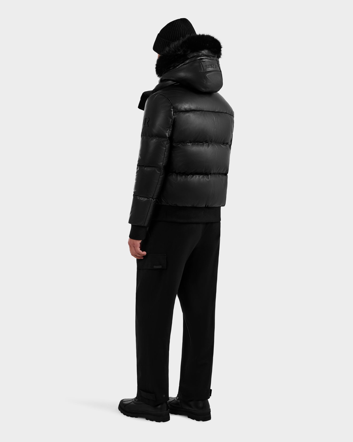 A person stands facing away, wearing a RUDSAK BOND X - 6122525 BLACK men’s winter jacket with RDS-certified down fill, black pants, beanie, and shoes against a plain light gray background.
