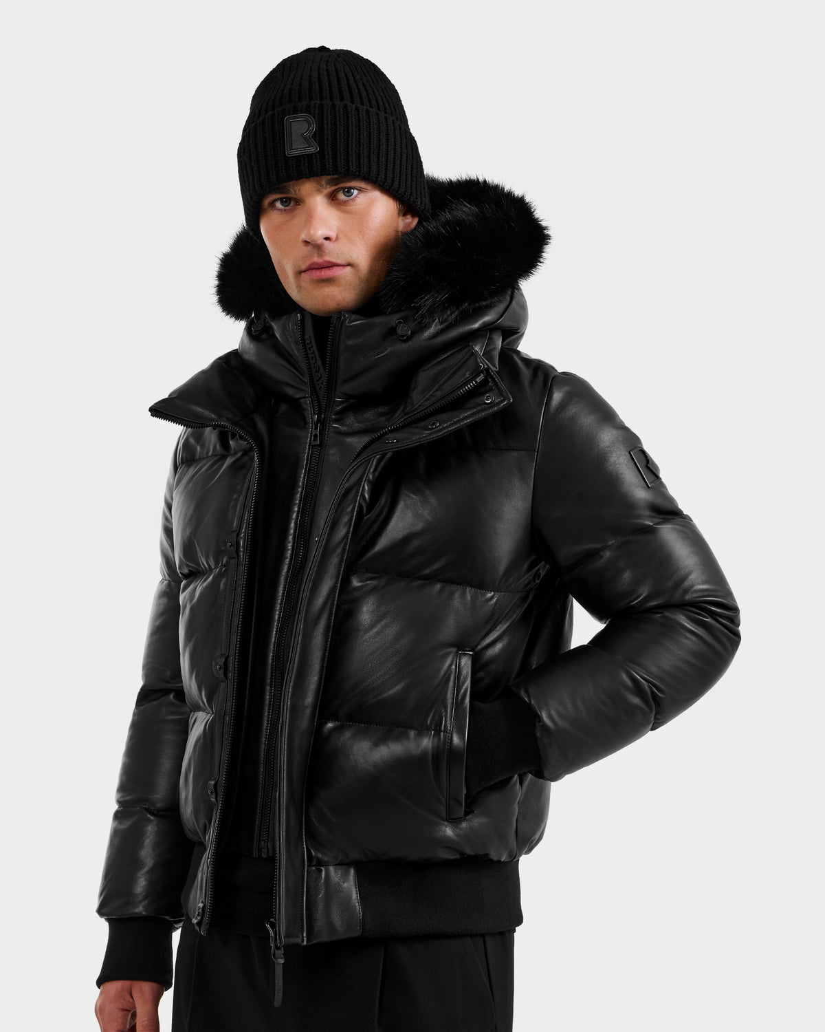 A person wearing a black beanie and the RUDSAK BOND X - 6122525 BLACK men’s winter jacket with RDS-certified down fill and a furry hood stands against a plain light background, looking forward with one hand in their pocket.