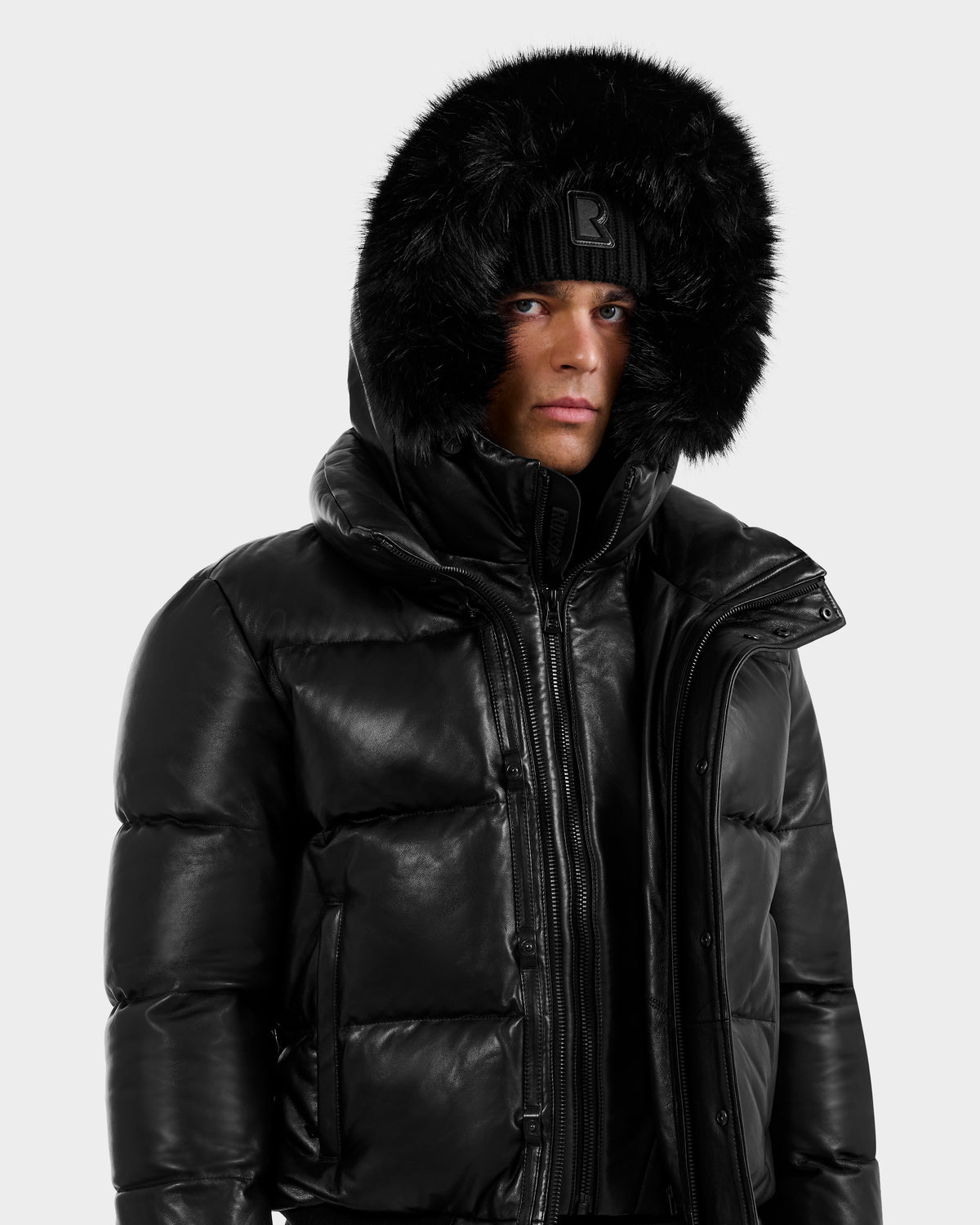 A person wearing the RUDSAK BOND X - 6122525 BLACK puffer jacket with RDS-certified down and a fur-lined hood, paired with a black beanie, looks forward neutrally against a plain, light background.