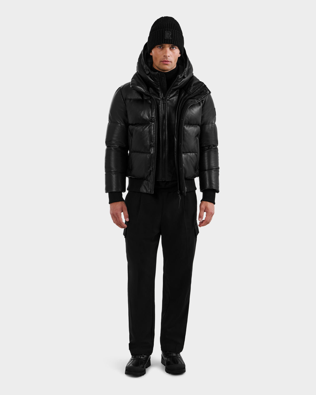 A man stands facing forward against a plain light background, wearing the RUDSAK BOND X - 6122525 BLACK men's winter jacket with RDS-certified down fill, layered over a black hoodie, paired with black pants, shoes, and a knit beanie.