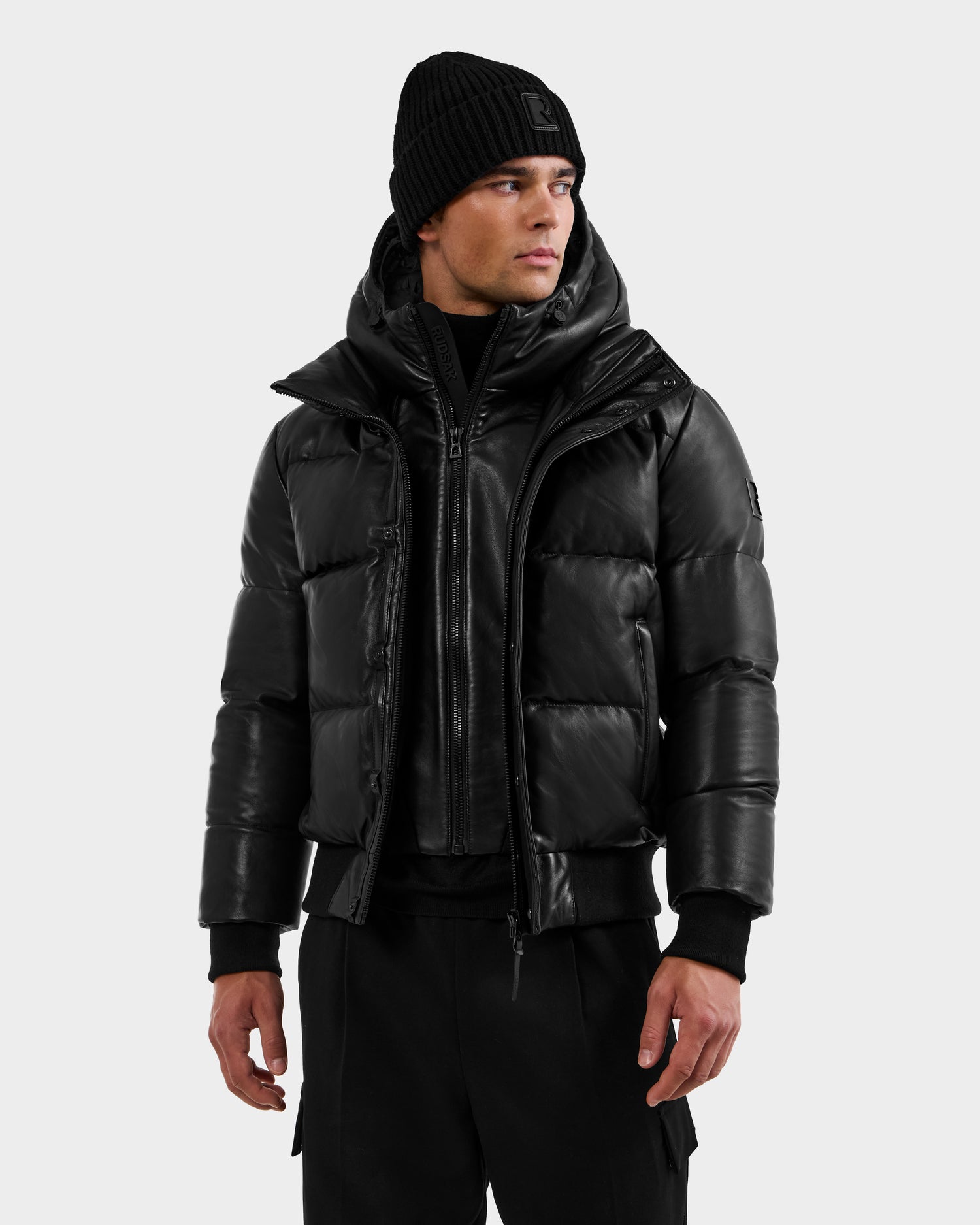 A man wearing the RUDSAK BOND X - 6122525 BLACK winter jacket, styled with a black beanie and black pants, stands against a plain light gray background, looking slightly to his left.