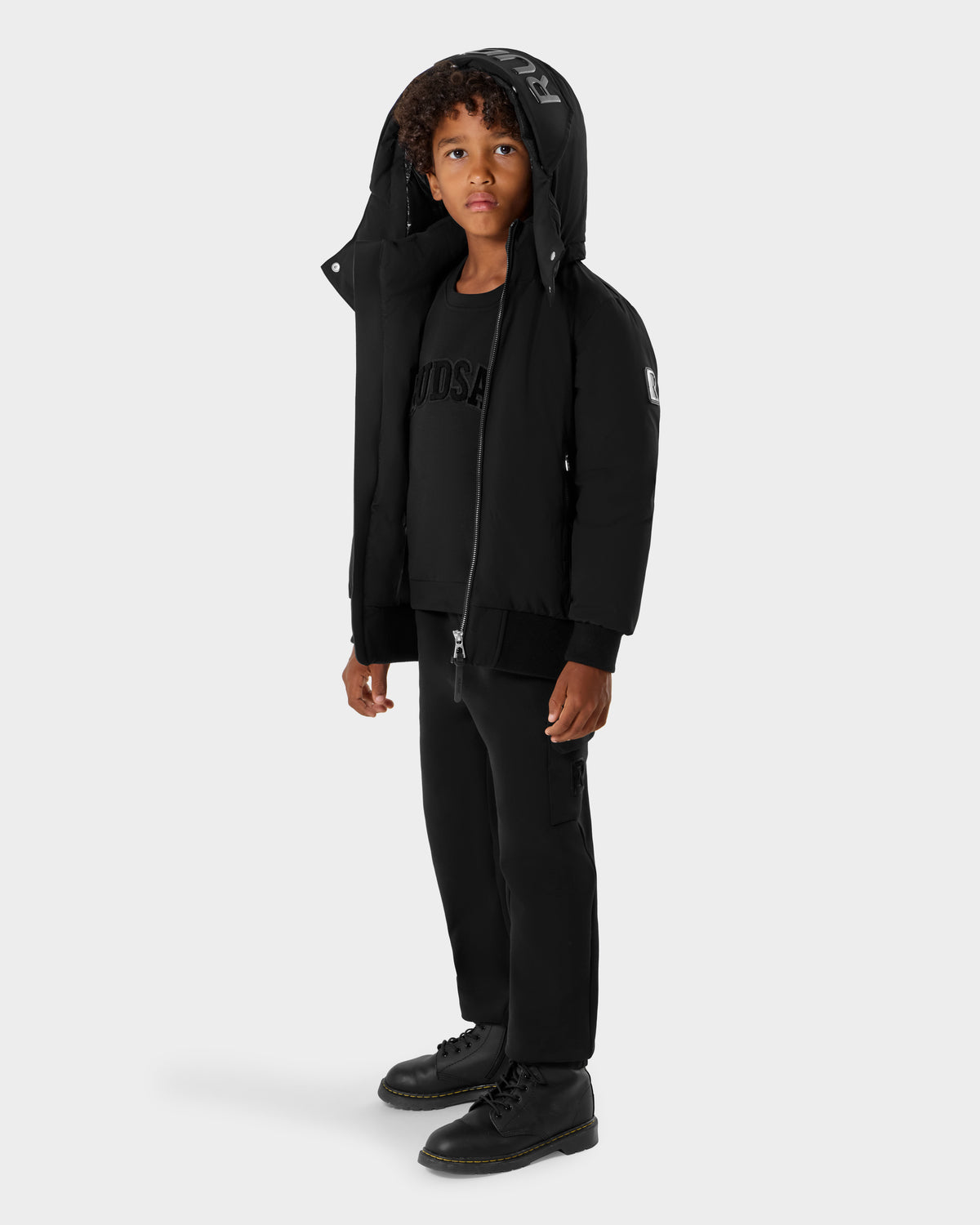 A young boy with curly hair wears the Rudsak BOOKER K - 0124500 Black weather-resistant kids' quilted puffer jacket with a removable hood, paired with black pants and boots, standing against a plain light gray background.