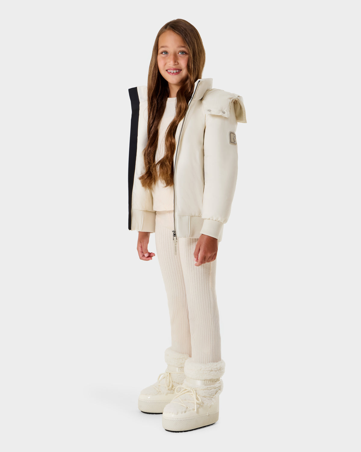 A young girl with long brown hair smiles in a Rudsak BOOKER K - 0124500 Vanilla kids' quilted puffer jacket with removable hood, matching sweater, ribbed pants, and chunky winter boots, against a plain white background.