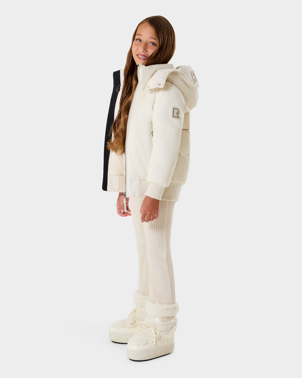 A young girl with long brown hair is wearing the Rudsak BOOKER K - 0124500 Vanilla kids' quilted puffer jacket with a removable hood, standing and smiling against a plain white background.
