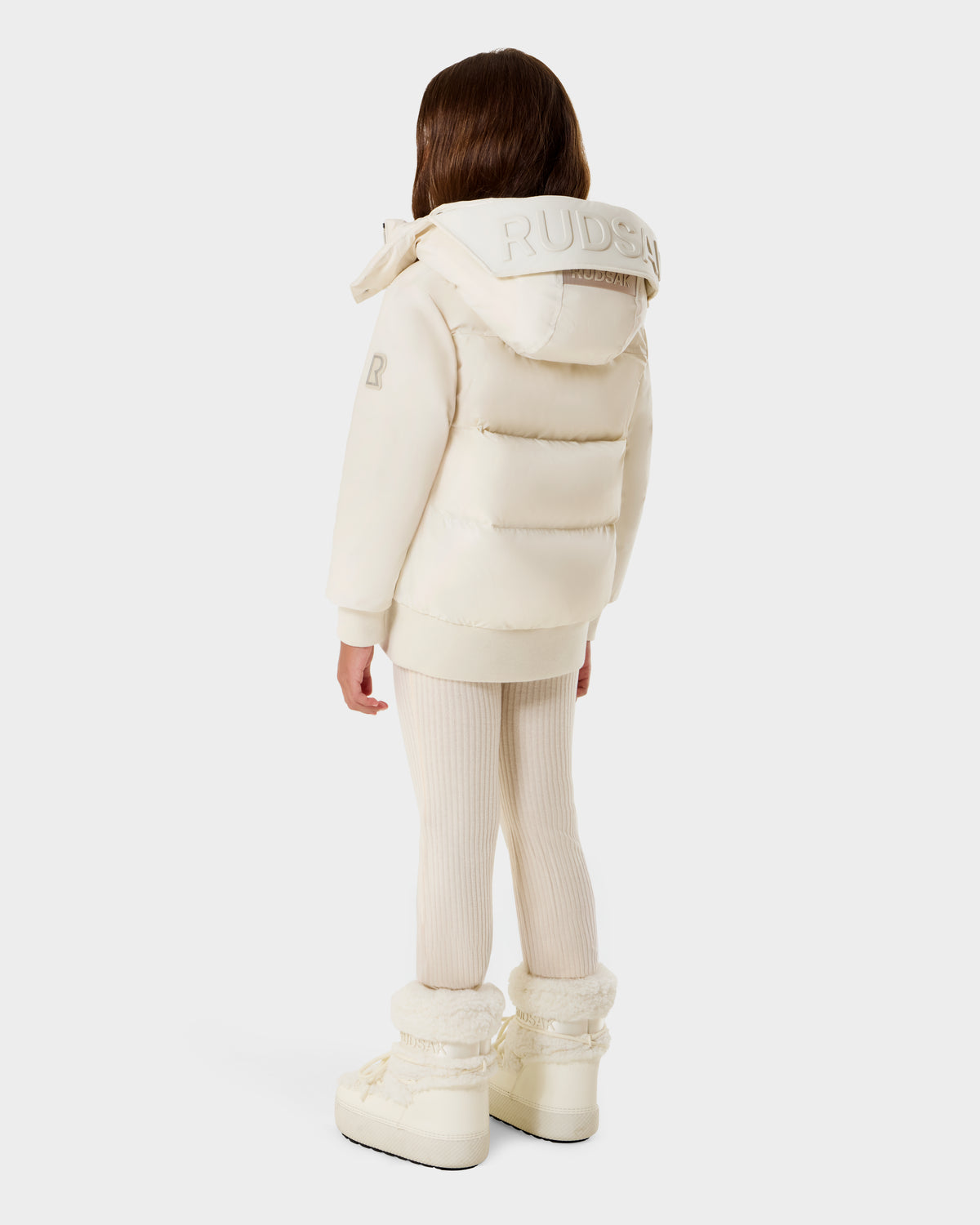 A young child with long brown hair, seen from behind, wears the Rudsak BOOKER K - 0124500 Vanilla kids’ quilted puffer jacket with a weather-resistant, removable hood, matching leggings, and cream winter boots against a plain light background.