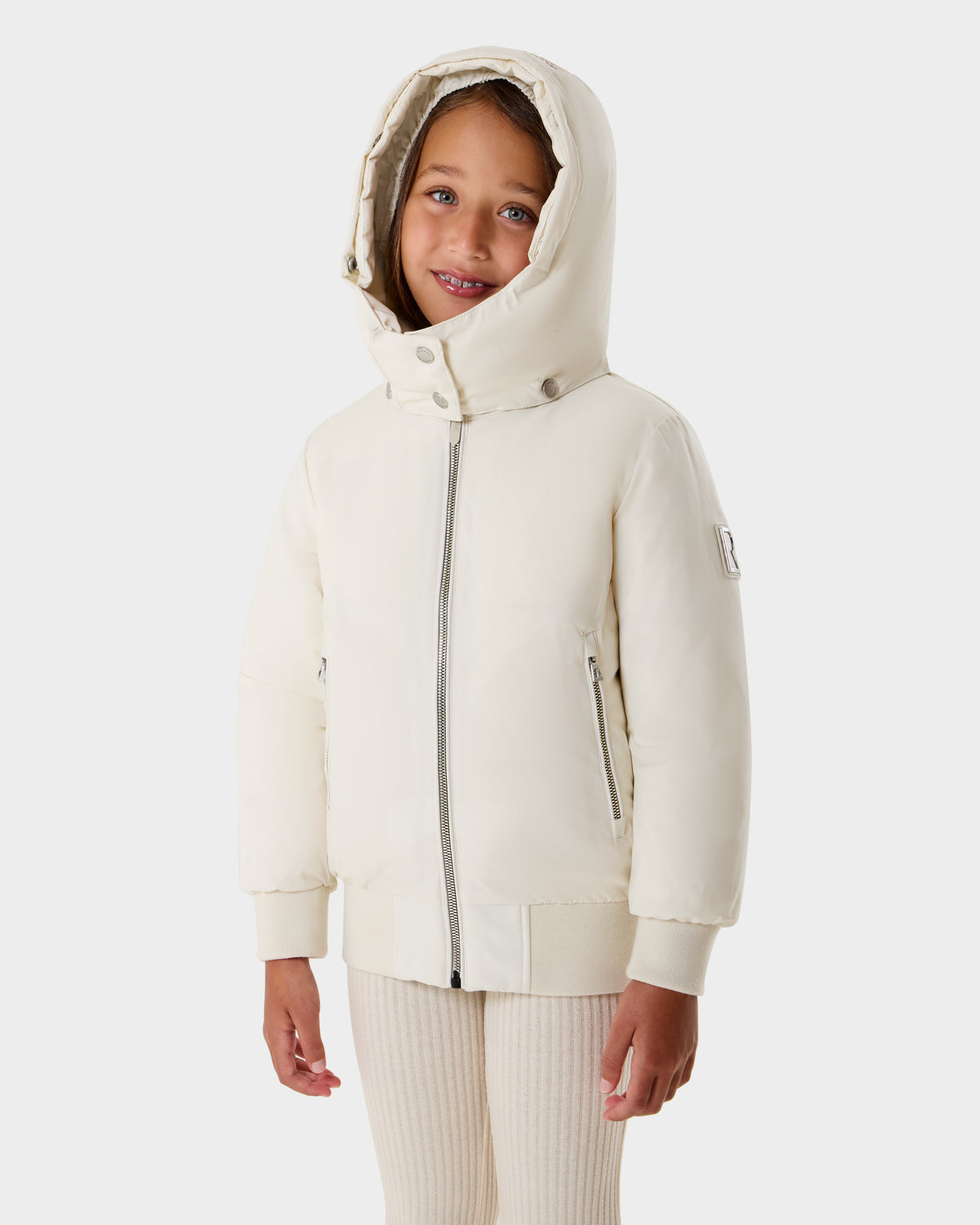 A young girl smiles in the Rudsak BOOKER K - 0124500 Vanilla kids' quilted puffer jacket with zipper, ribbed cuffs, and a removable hood, standing against a plain white background.