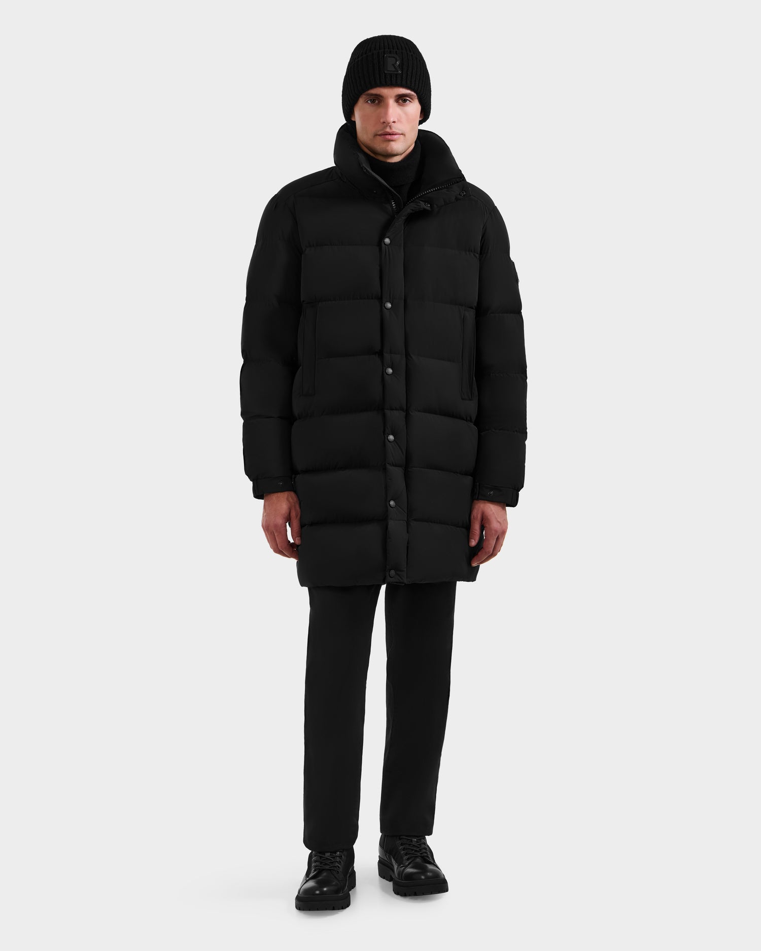 A person stands facing forward, wearing Rudsak’s BRAYDEN M-M - M125535 Black men’s down puffer coat, paired with a black beanie, pants, and shoes against a light gray background.
