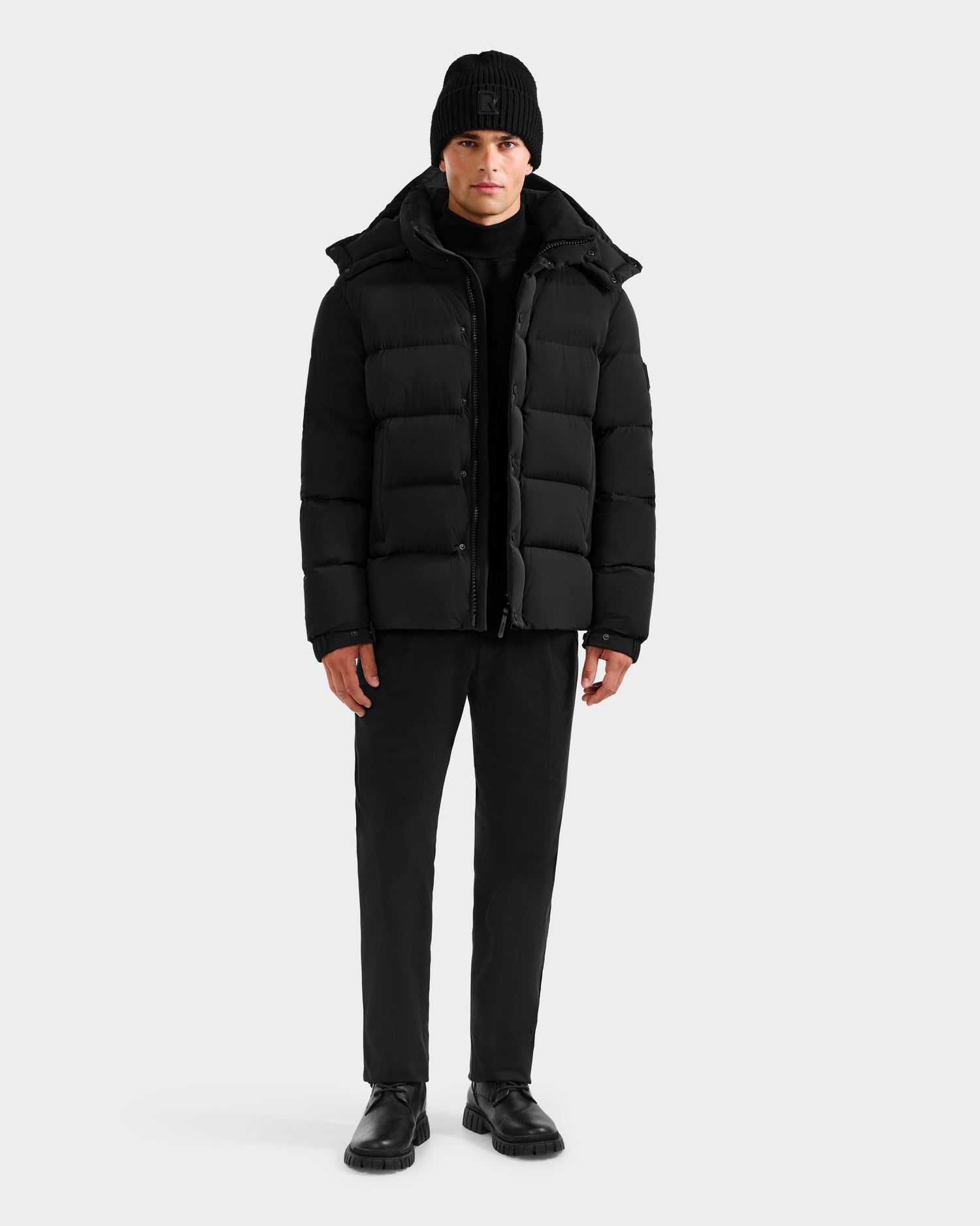 A person faces forward in a Rudsak BRAYDEN M-S - M125524 Black men’s down puffer jacket with RDS-certified down, paired with black pants, boots, and a black beanie against a plain light gray background.