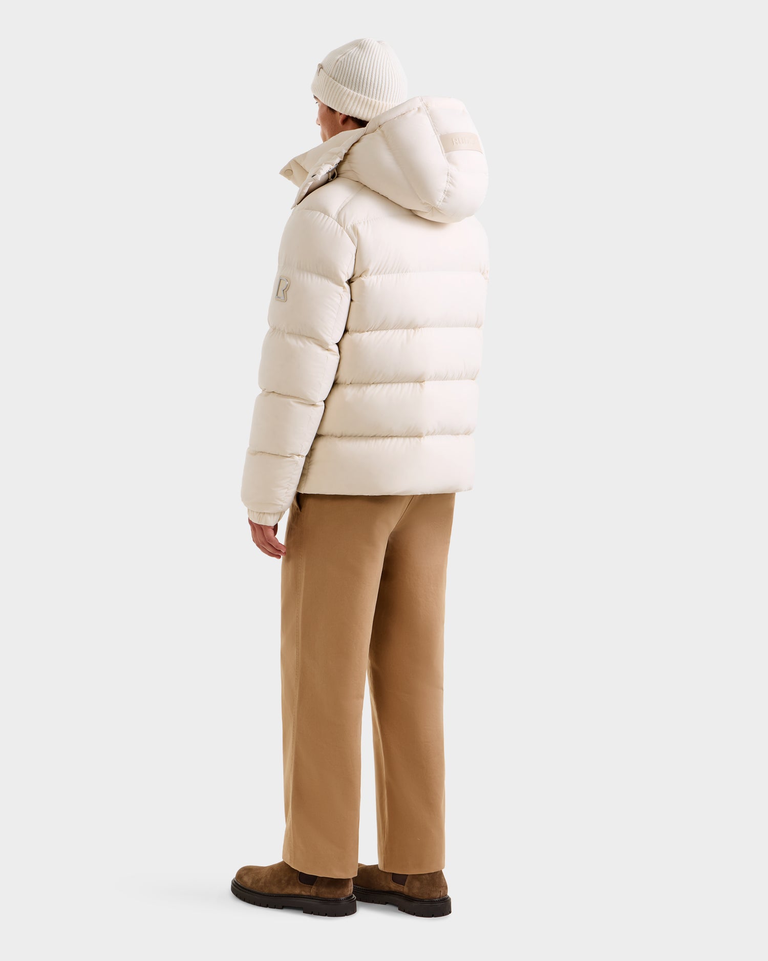 A person stands facing away, wearing the Rudsak BRAYDEN M-S - M125524 Vanilla jacket with a hood, beige pants, brown shoes, and a white knit beanie against a plain light background.