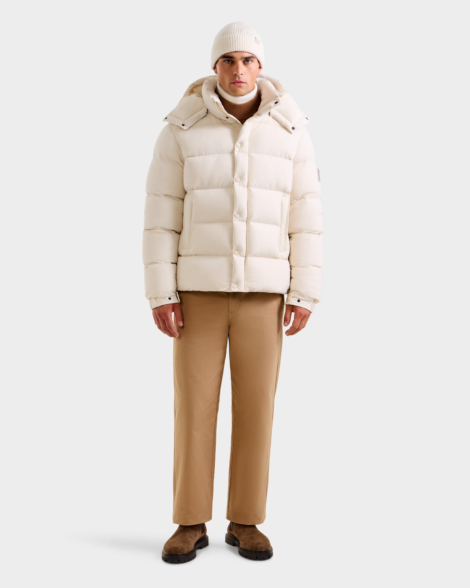 A man stands facing forward against a plain light background, wearing the Rudsak BRAYDEN M-S - M125524 Vanilla puffer jacket, a beige knit beanie, tan pants, and brown shoes.