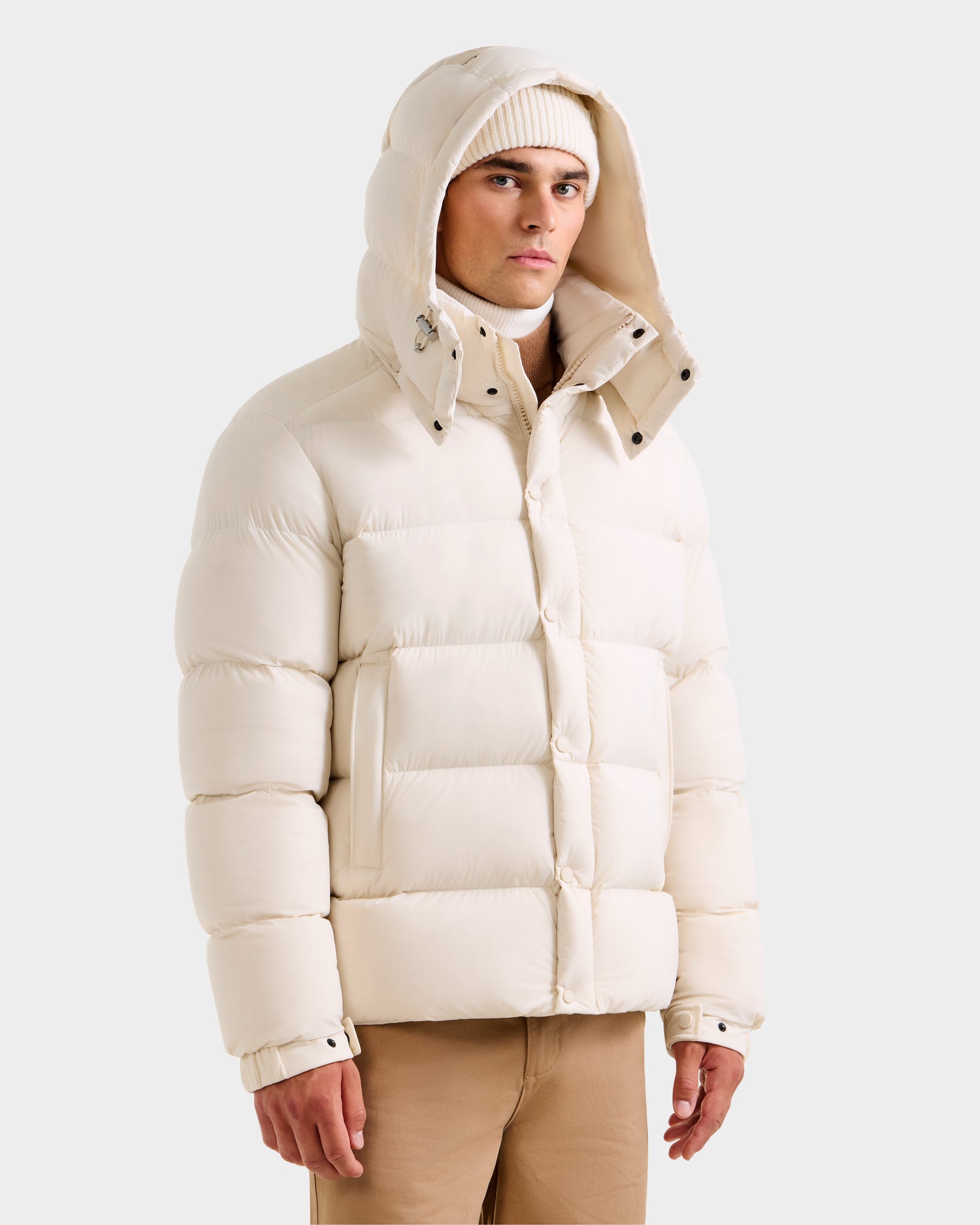 A man stands against a plain light background wearing the Rudsak BRAYDEN M-S - M125524 Vanilla men’s down puffer jacket with hood, a matching knit beanie, light tan pants, and a white turtleneck for a stylish winter look.