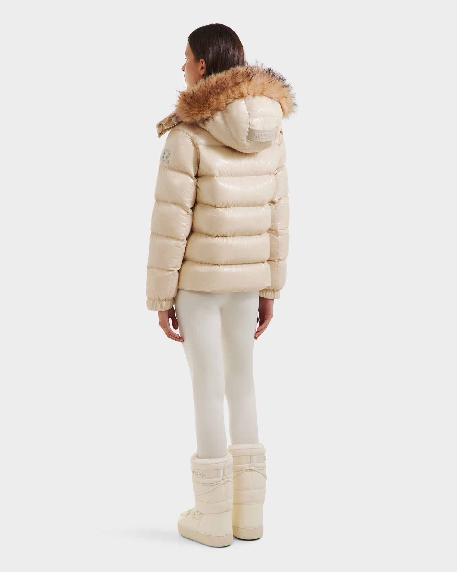 A woman faces away, wearing the Rudsak BREE S - L125583 Vanilla women's down puffer jacket with ultra-gloss finish and fur-trimmed hood, paired with cream leggings and matching winter boots against a plain white background.