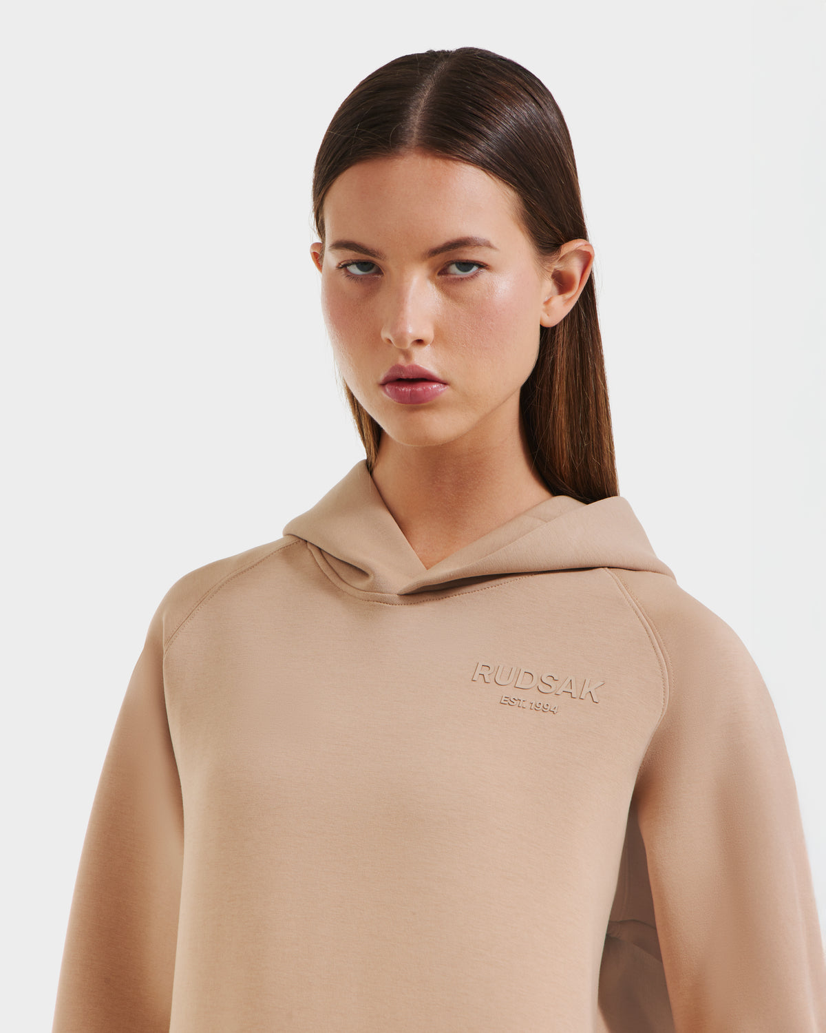 A woman with straight brown hair wears the Rudsak BRIAR - 9525001 Almond W beige unisex premium cotton-blend hoodie, featuring RUDSAK embroidered on the chest, standing against a plain white background and looking at the camera.