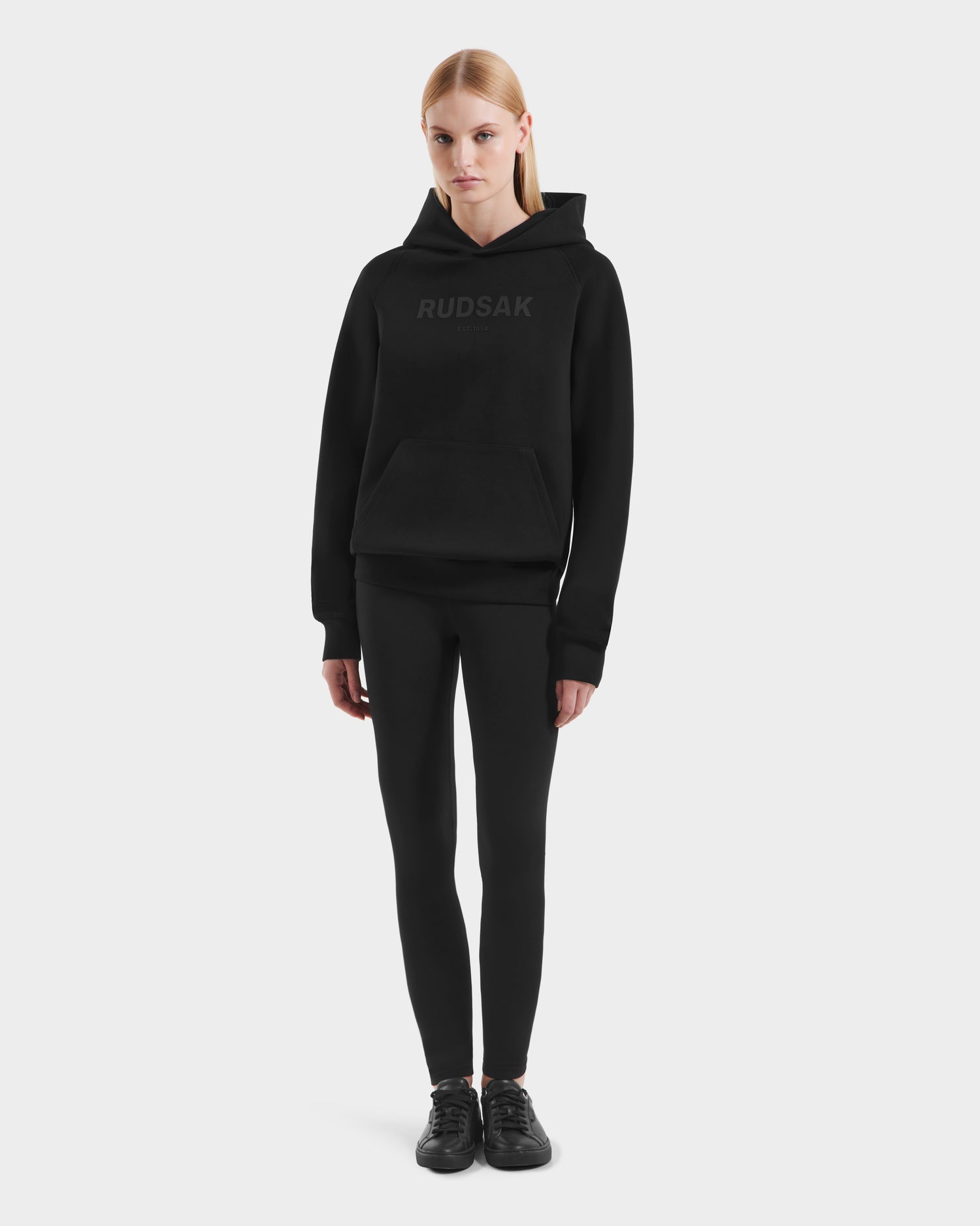 A woman with straight blonde hair models the Rudsak BRIAR - 9525001 Black W unisex hoodie against a white background, paired with black leggings and sneakers. She faces forward with a neutral expression, showcasing a casual look.