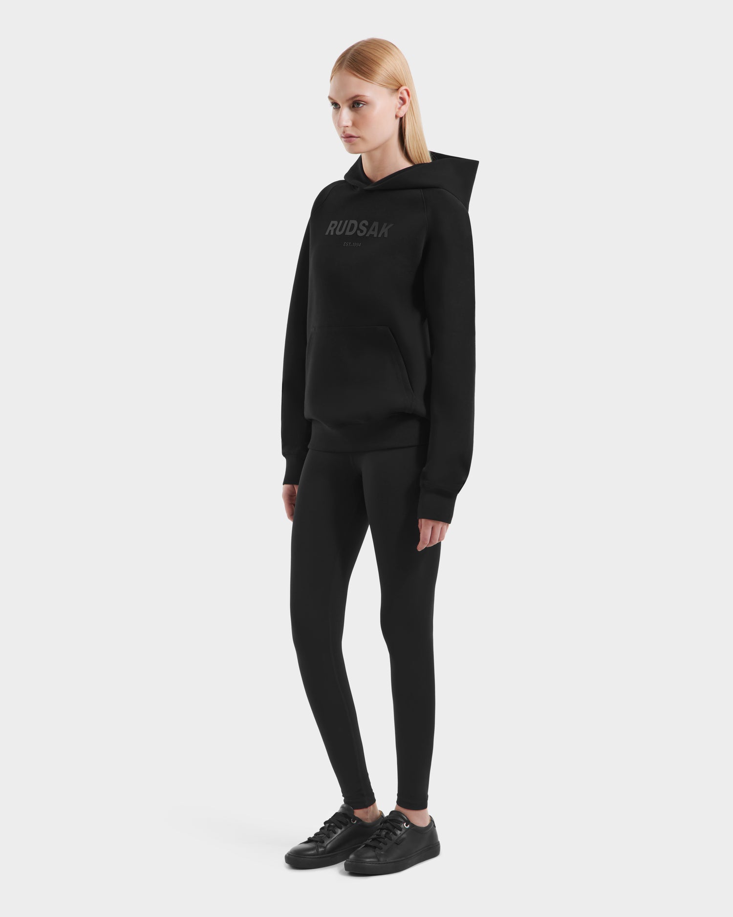 A woman with straight blonde hair models the Rudsak BRIAR - 9525001 Black W hoodie against a white background, styled with black leggings and sneakers for a casual look.