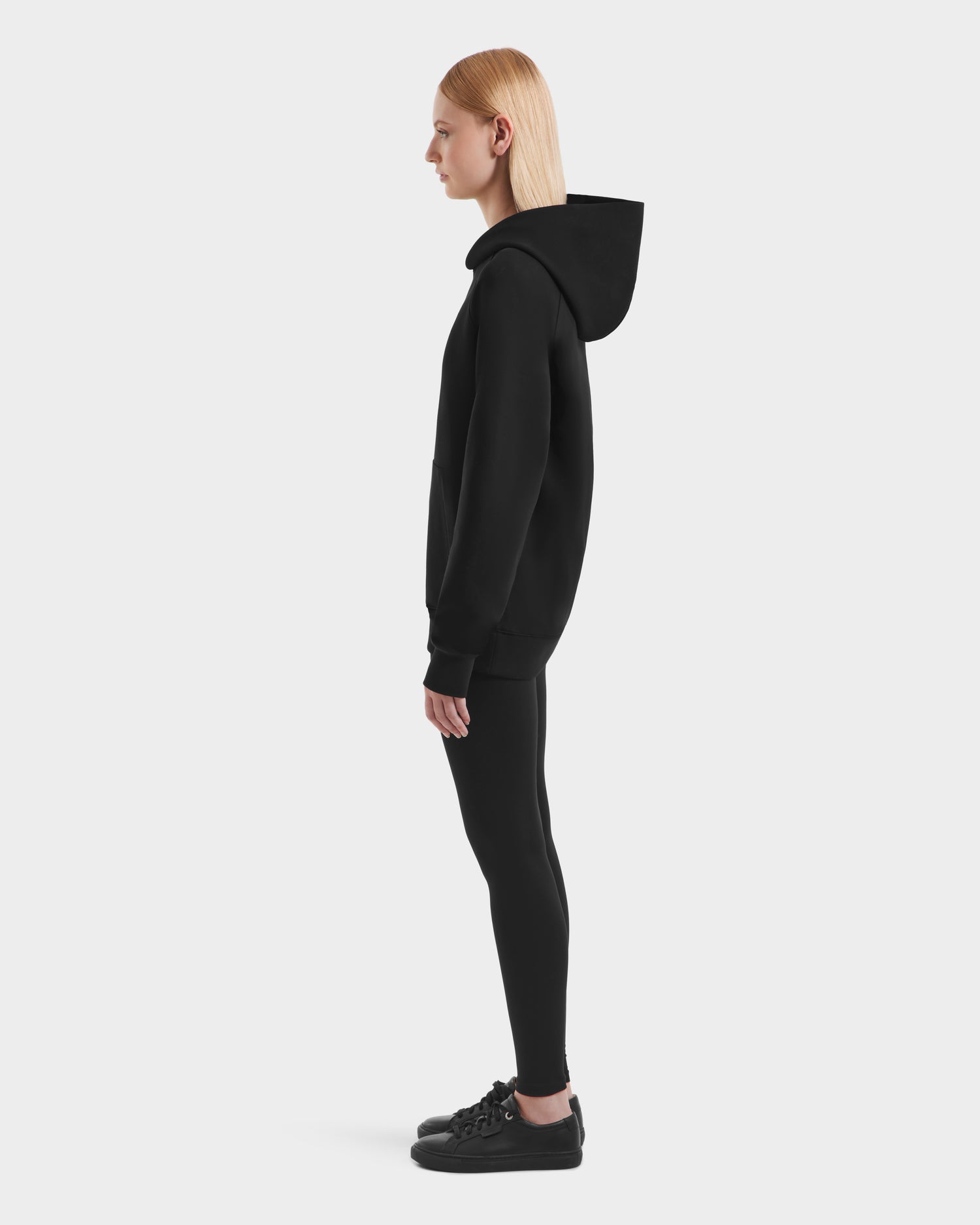 A person with straight blonde hair stands in profile, wearing the Rudsak BRIAR - 9525001 Black W hoodie with black leggings and black sneakers against a white background for a clean, casual look.