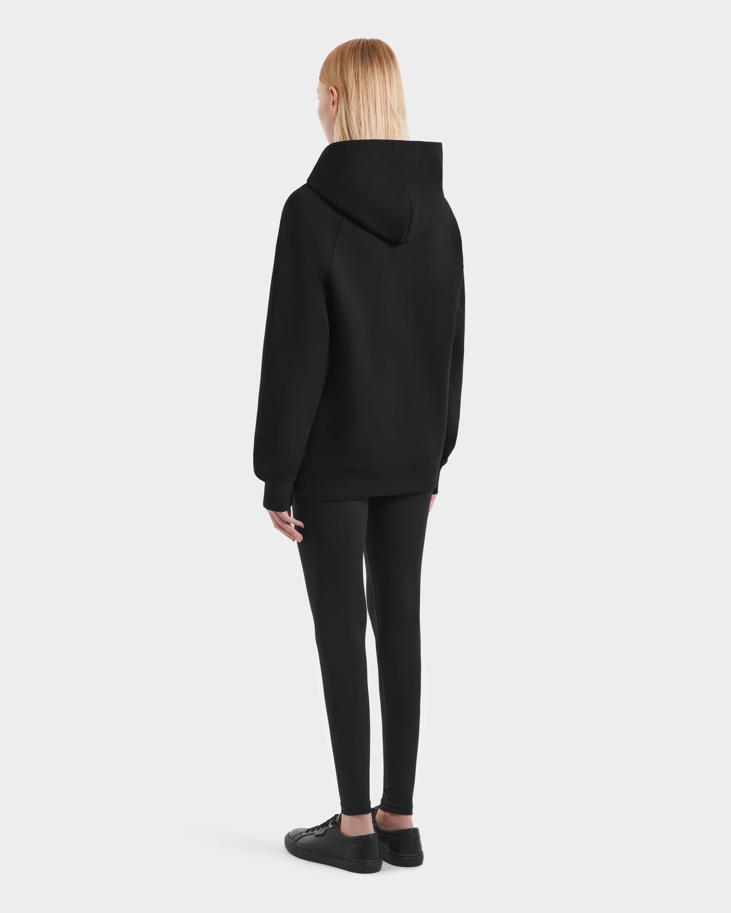 A person with straight blond hair faces away, wearing the Rudsak BRIAR - 9525001 Black W unisex hoodie, black leggings, and black sneakers for a casual look against a plain white background.