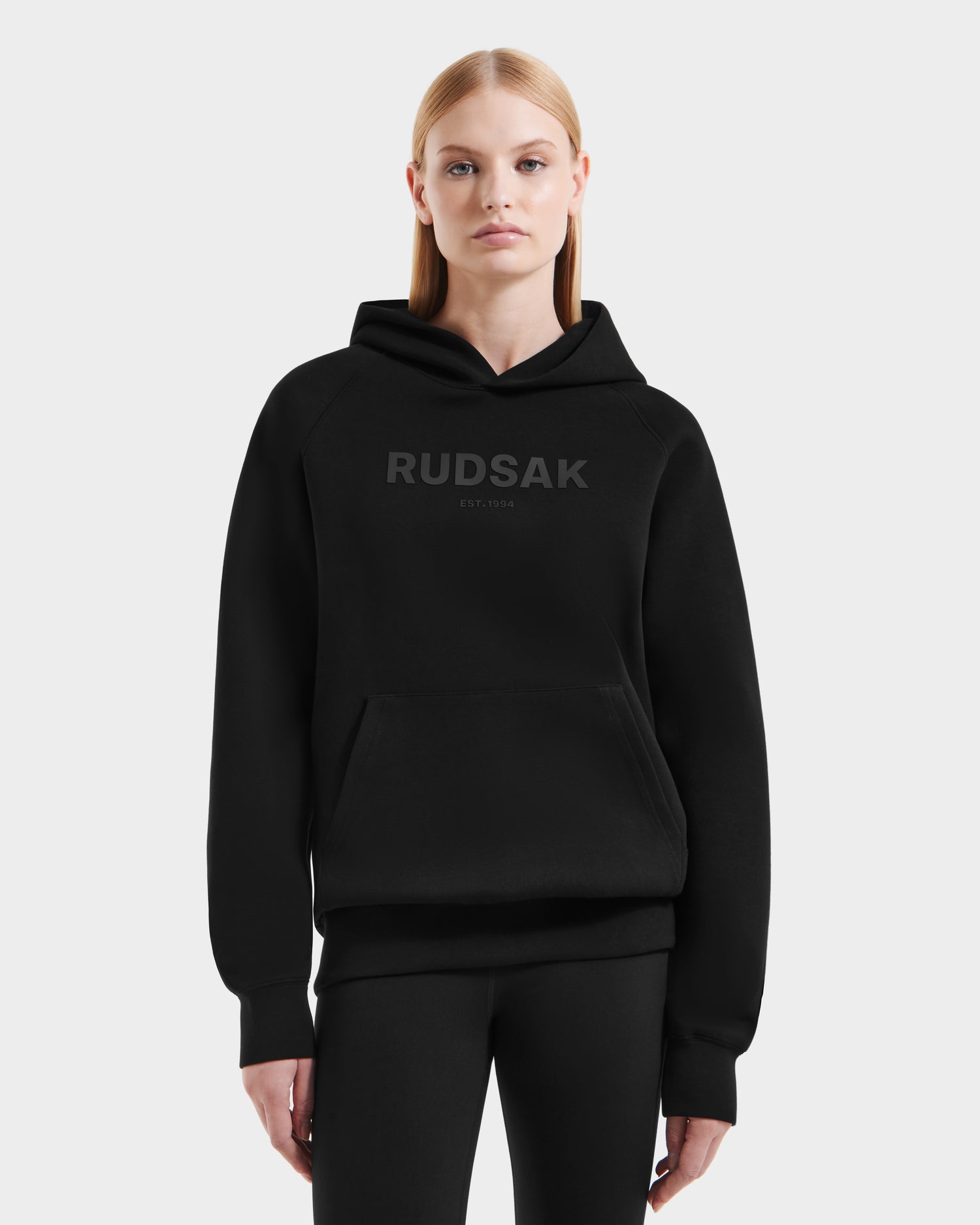A woman with straight blonde hair wears the Rudsak BRIAR - 9525001 Black W hoodie, made from a premium cotton blend with "RUDSAK EST. 1994" on the front, paired with black leggings against a white background.