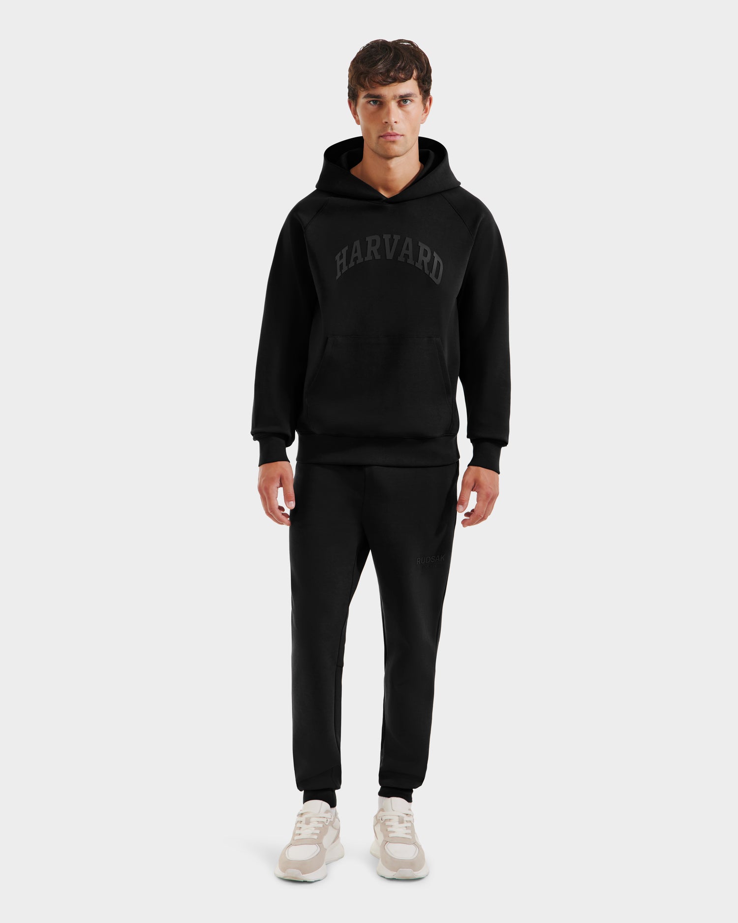 A man stands facing forward in a limited-edition Harvard THE COOP hoodie, Rudsak BRIAR R HV - H525506 Black jogger pants, and white sneakers against a plain, light-colored background.