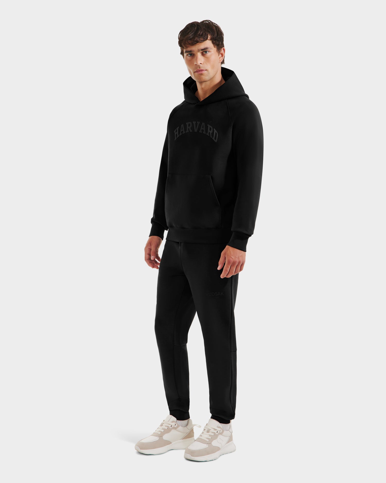 A man stands against a plain light gray background wearing Rudsak's BRIAR R HV - H525506 Black hoodie with HARVARD on the front, paired with black joggers and white sneakers.
