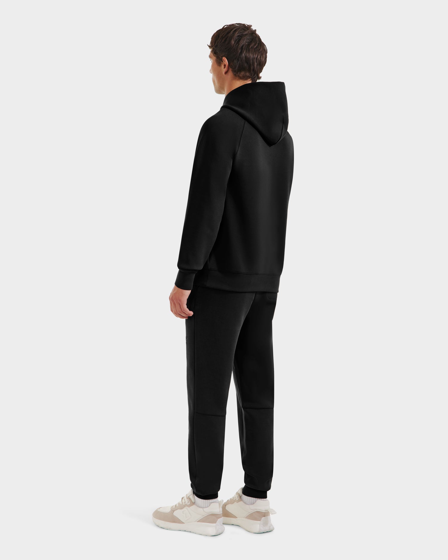 A person wearing the Rudsak BRIAR R HV - H525506 Black hoodie and matching black sweatpants stands facing away with their head turned, paired with light sneakers, all set against a plain white background.